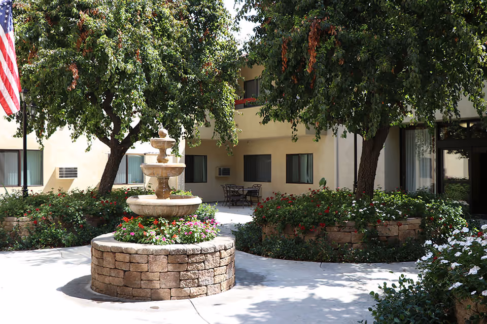 A landscaped courtyard with a stone tiered fountain surrounded by flowers, trees, and outdoor seating in front of a beige senior living building.