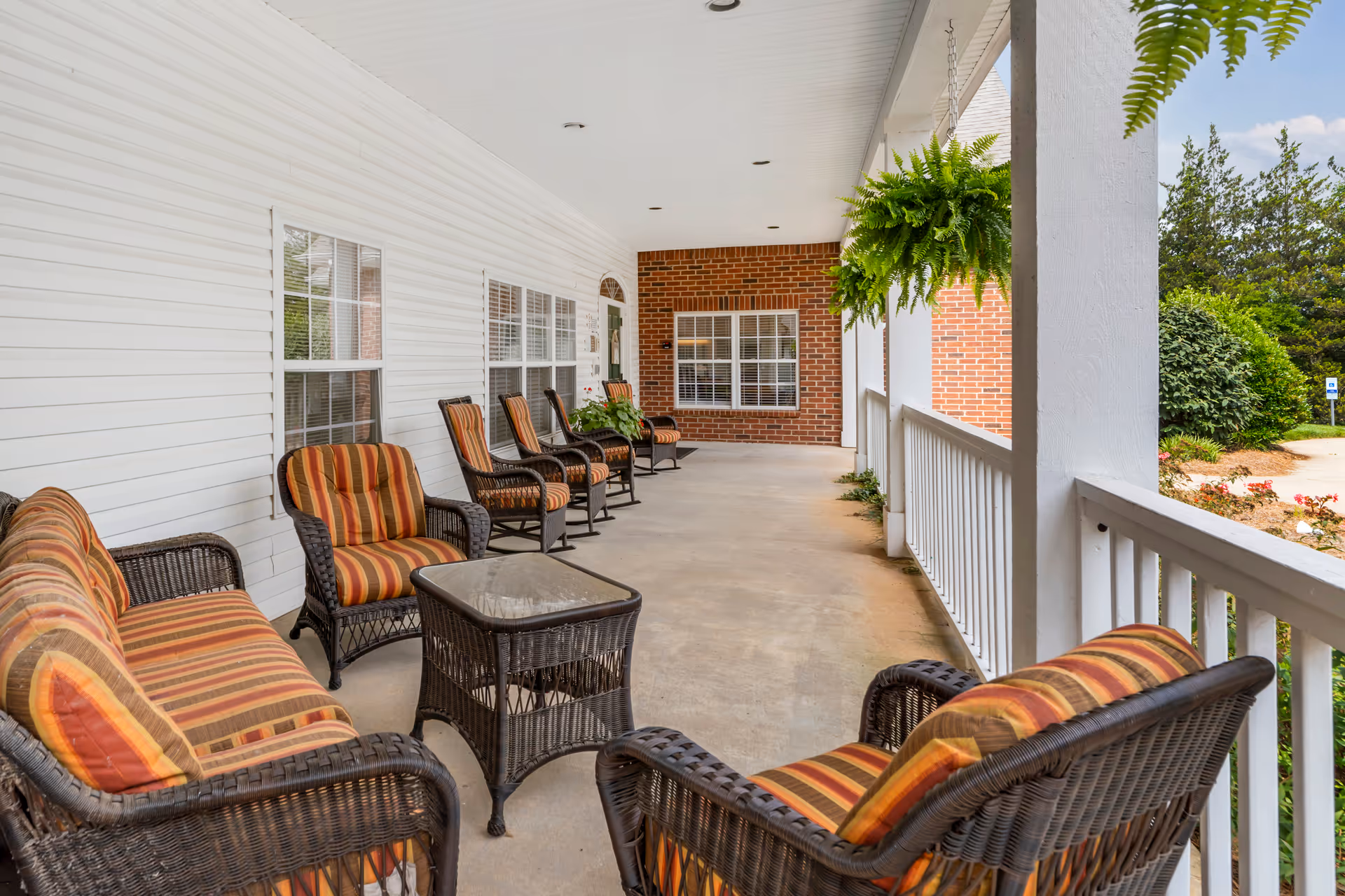 Covered outdoor porch area with wicker chairs and sofas featuring striped cushions arranged along the wall and railing. Hanging green plants are suspended from the ceiling, and there is a brick wall with windows at the far end. The porch overlooks a landscaped garden area with bushes and flowers.