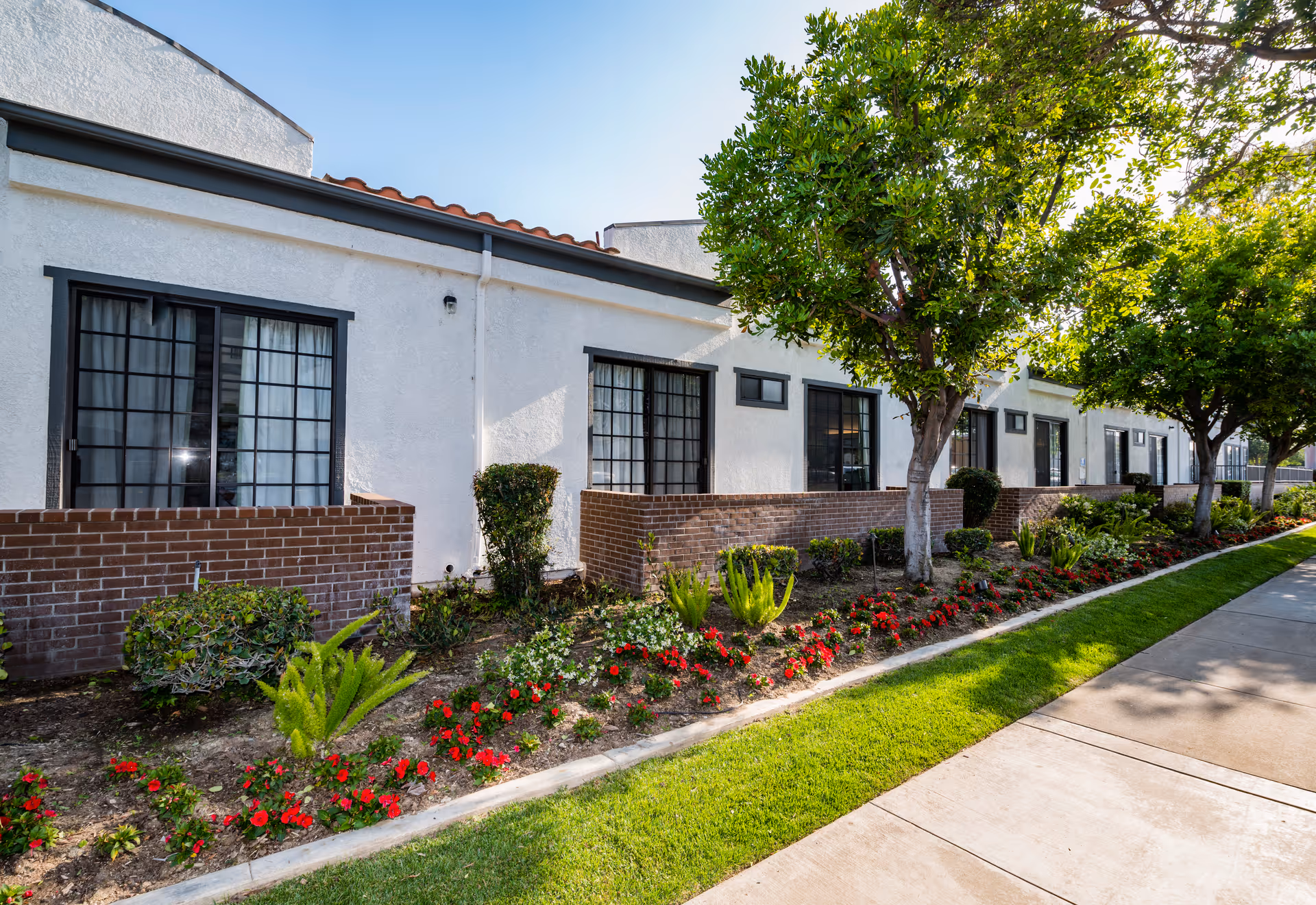 Front exterior of a white single-story building with brick planter walls, landscaped flowerbeds, trees, and a sidewalk.