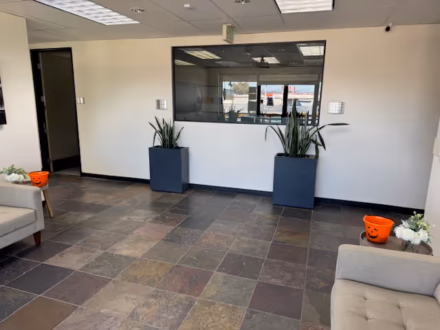 Indoor lobby with tiled floor, seating, and tall planters by a windowed service area.