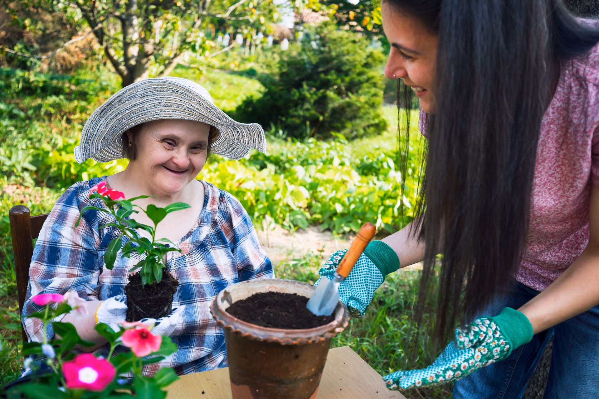 Two women garden together outdoors, one elderly resident wearing a hat, with another helping pot flowers