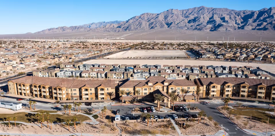 Aerial view of a multi-story retirement community building with parking, palm trees, surrounding homes, and mountains in the background.
