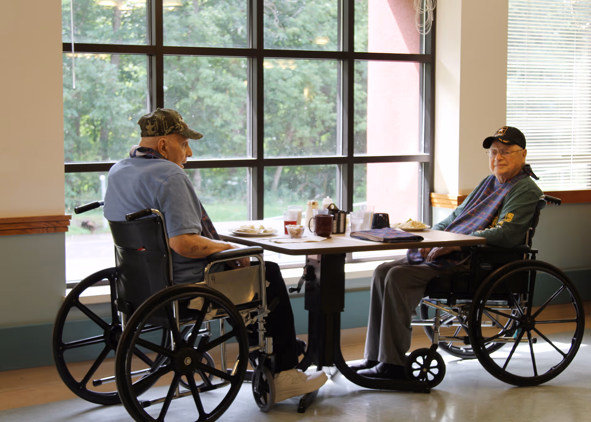 Two elderly men in wheelchairs sit at a dining table by large windows in a senior living facility.