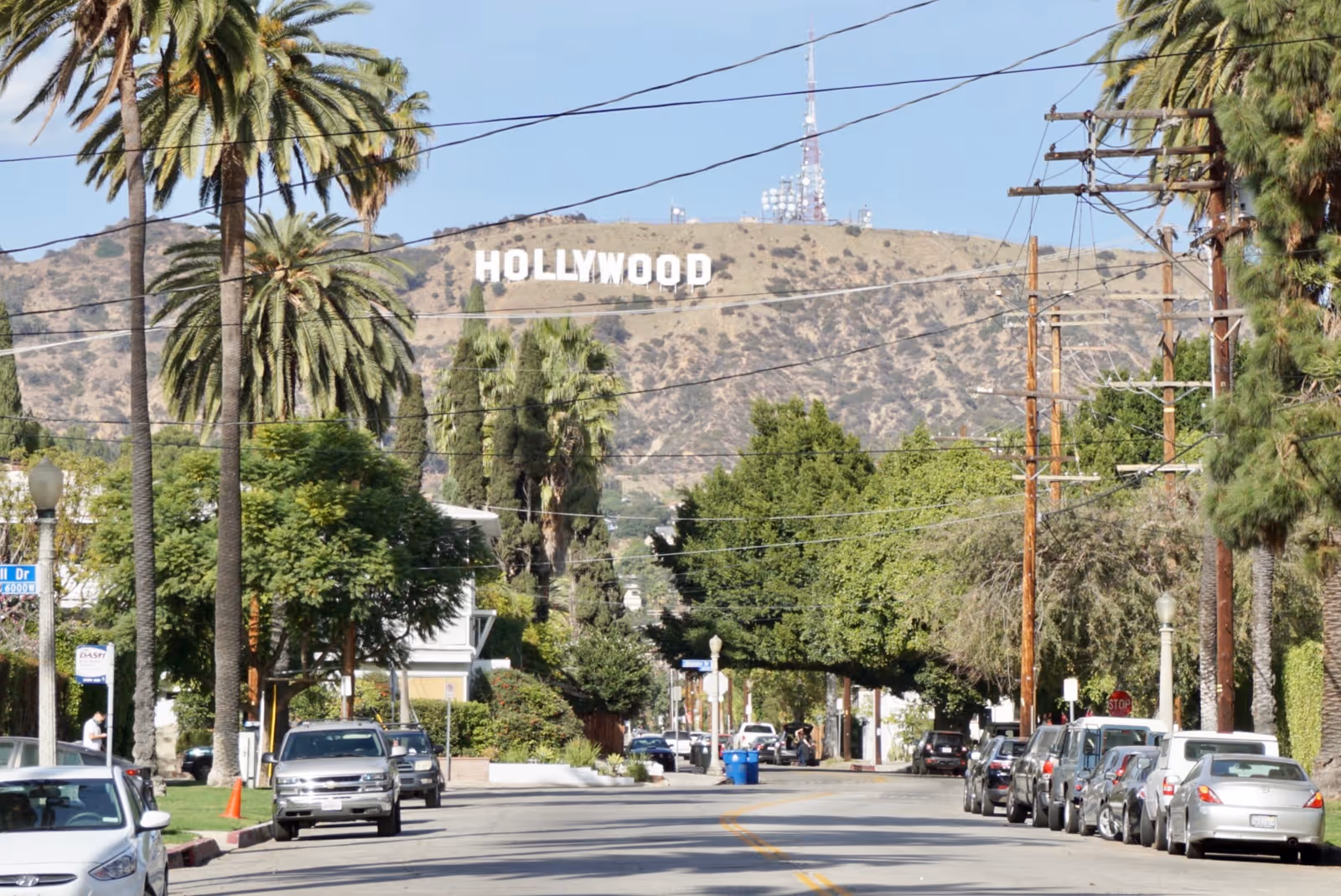 Wide street lined with palm trees and parked cars, with the Hollywood sign on a hillside.