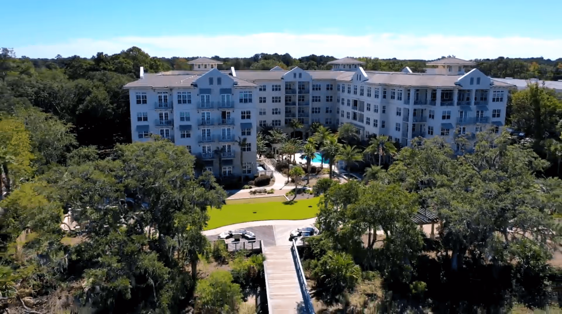 A large, multi-story residential building surrounded by lush green trees and landscaping. The building has multiple balconies and windows, with a swimming pool visible in the courtyard area. A wooden walkway leads up to the entrance of the building.