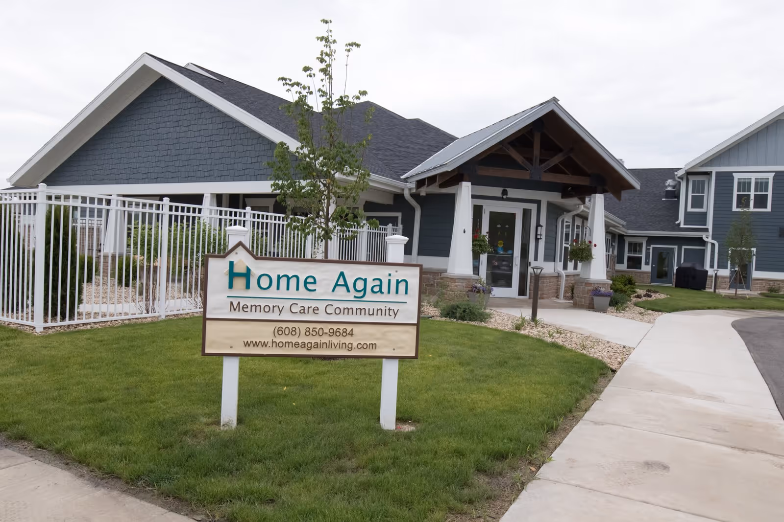 Exterior view of Home Again Assisted Living & Memory Care Waunakee building with a sign in front displaying the facility name, phone number, and website. The building has a blue-gray exterior with white trim, a covered entrance, and a fenced area to the left. There is a sidewalk leading to the entrance and well-maintained grass and landscaping around the building.