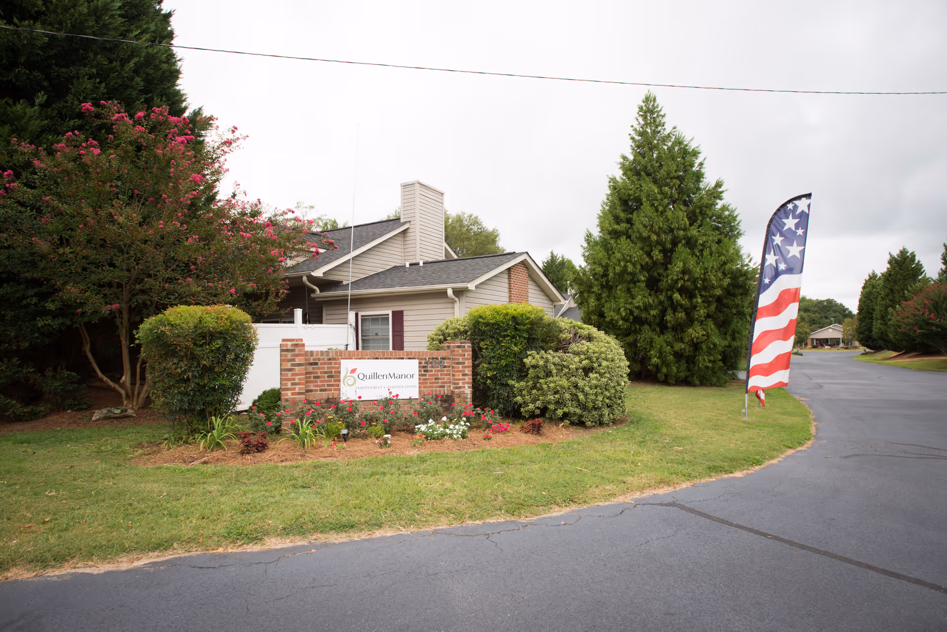Exterior view of Quillen Manor Independent and Assisted Living facility showing a brick sign with the facility's name surrounded by bushes and flowers. There is a paved driveway and an American flag banner on the right side. Trees and a building are visible in the background under a cloudy sky.