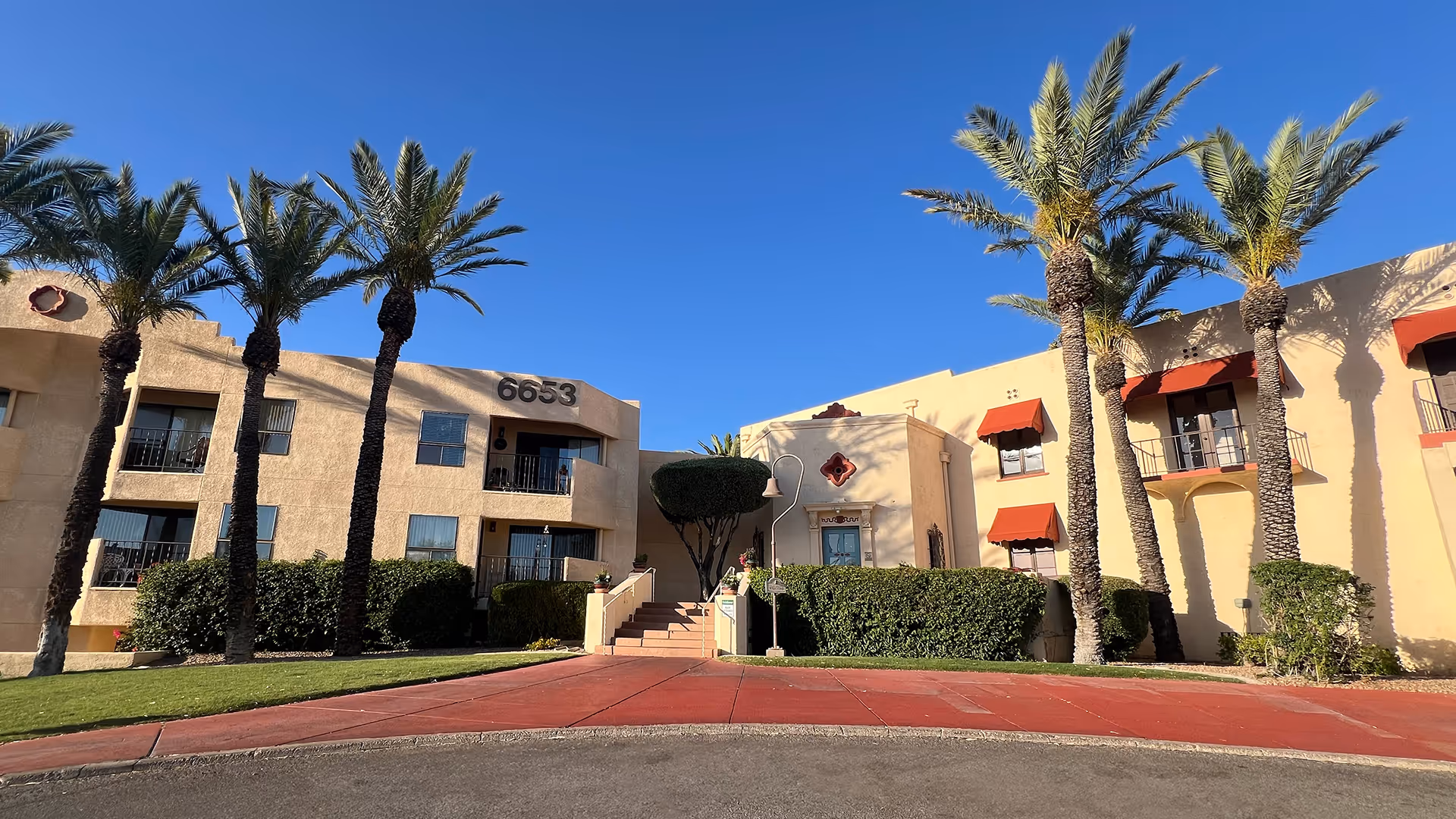 Atria Bell Court Gardens exterior with tan stucco buildings, palm trees, and red awnings.