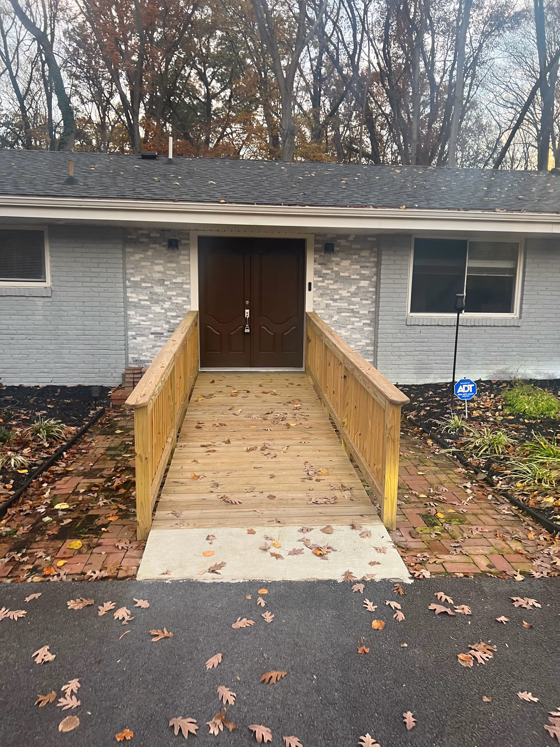 Front entrance of a single-story assisted living facility with wooden ramp and brown double doors.