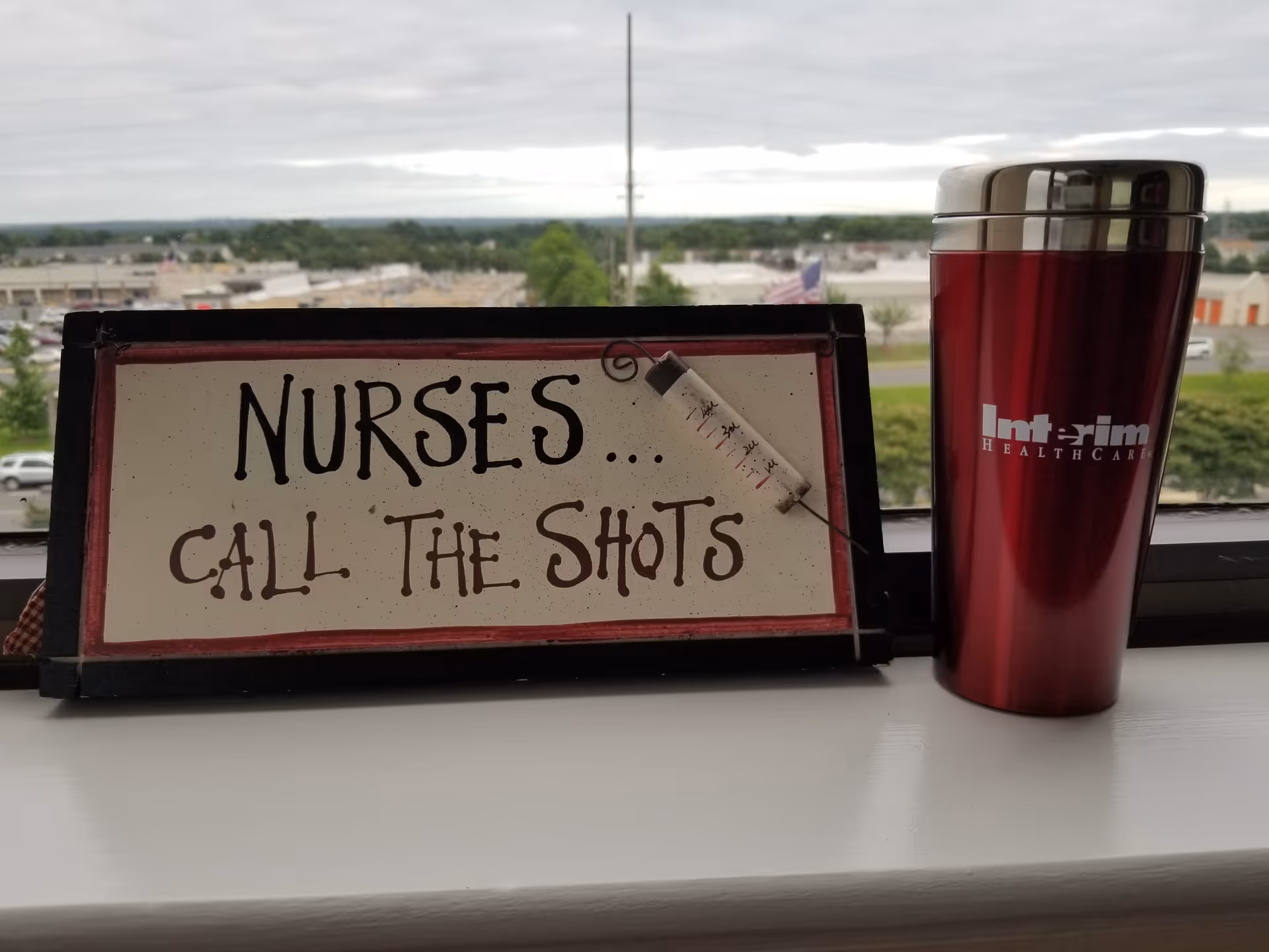 Sign reads 'NURSES... CALL THE SHOTS' beside a maroon travel mug on a windowsill.