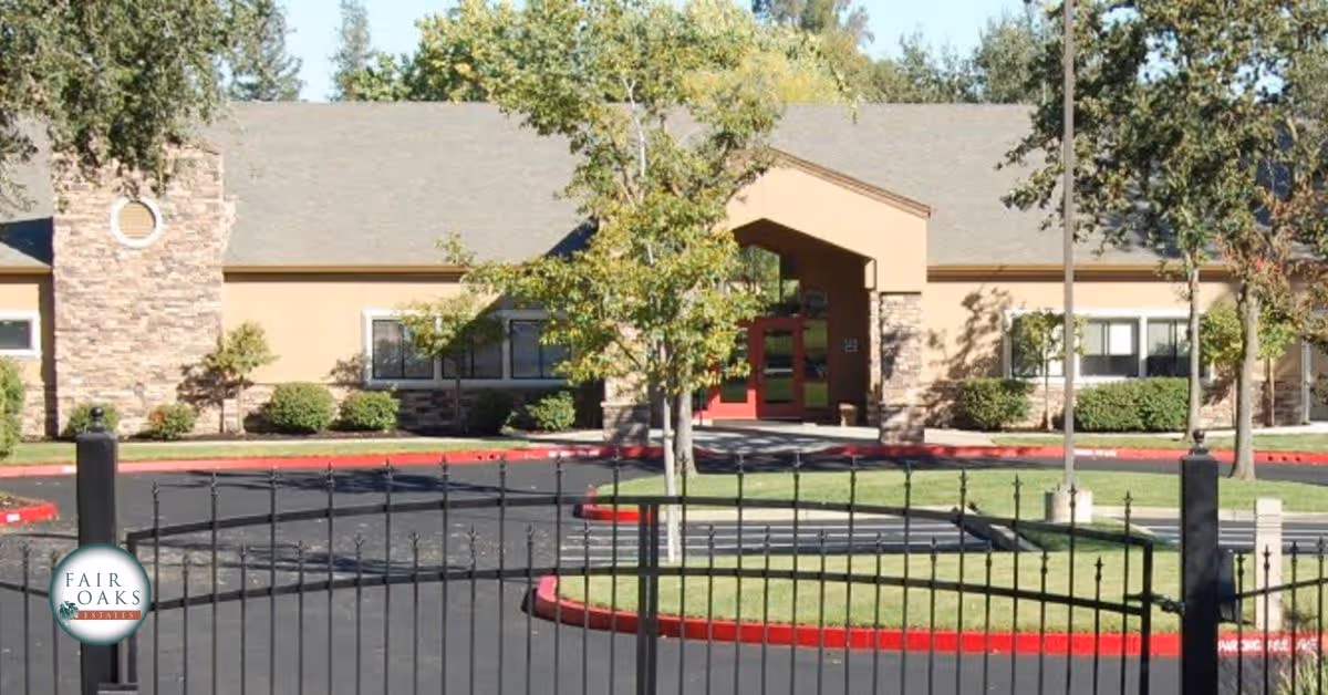 Exterior view of Fair Oaks Estates building with a gated entrance, a paved driveway, green trees, and shrubs around the building.