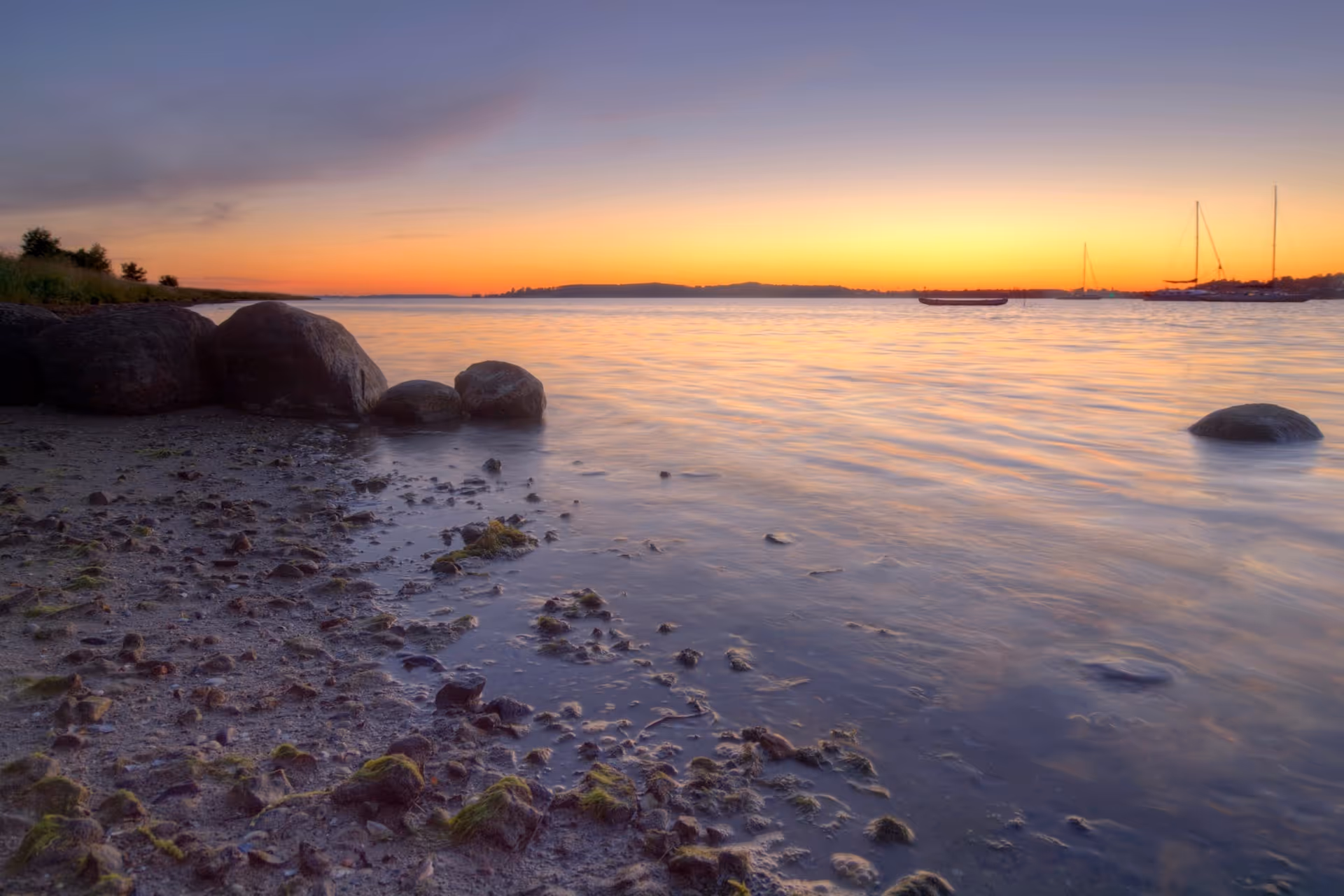 Sunset over a rocky shoreline with calm water and distant sailboats.