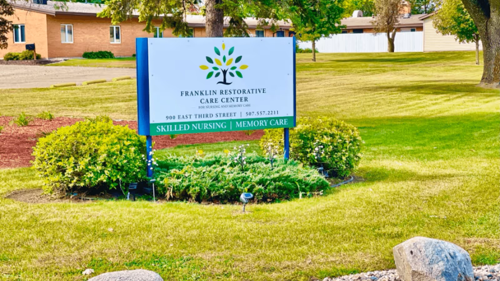 Outdoor view of a green lawn with bushes and trees surrounding a sign for Franklin Restorative Care Center. The sign includes the facility's name, address, phone number, and mentions skilled nursing and memory care services. A brick building is visible in the background.