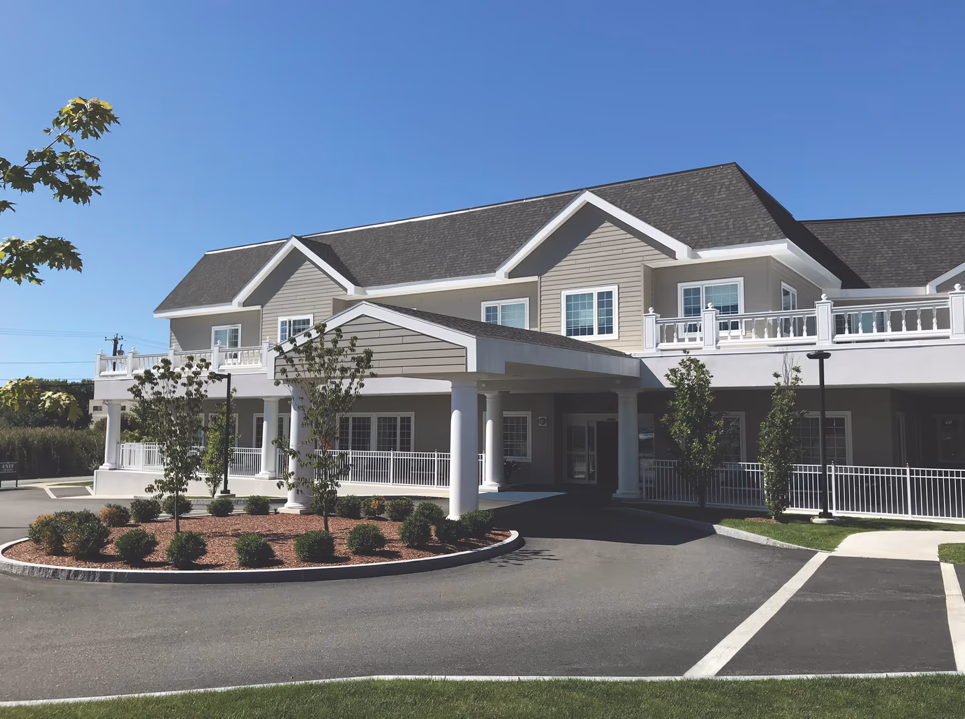 Exterior view of Port Healthcare Center, a two-story building with beige siding and white trim. The entrance features a covered drop-off area supported by white columns, surrounded by landscaped bushes and small trees under a clear blue sky.