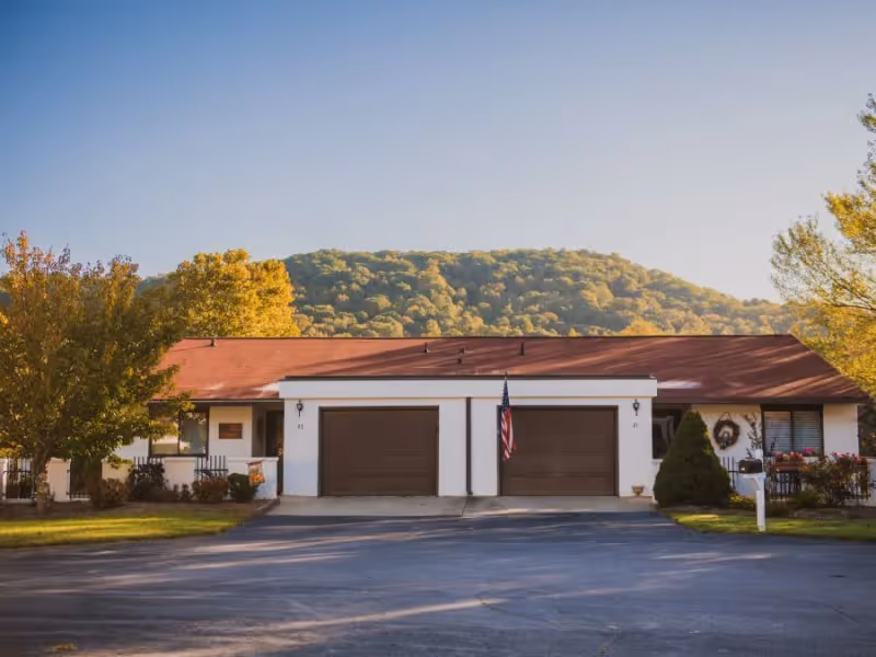 Single-story retirement community building with two garage doors and an American flag, set against a tree-covered hill.