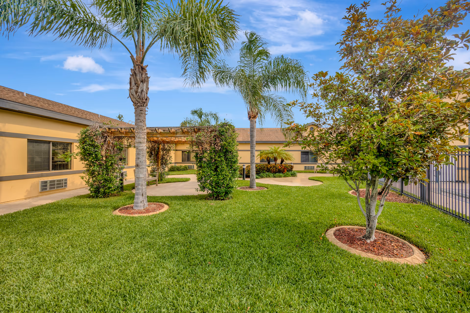 Sunny courtyard with palm trees, manicured lawn, and tan exterior buildings surrounding walkways.