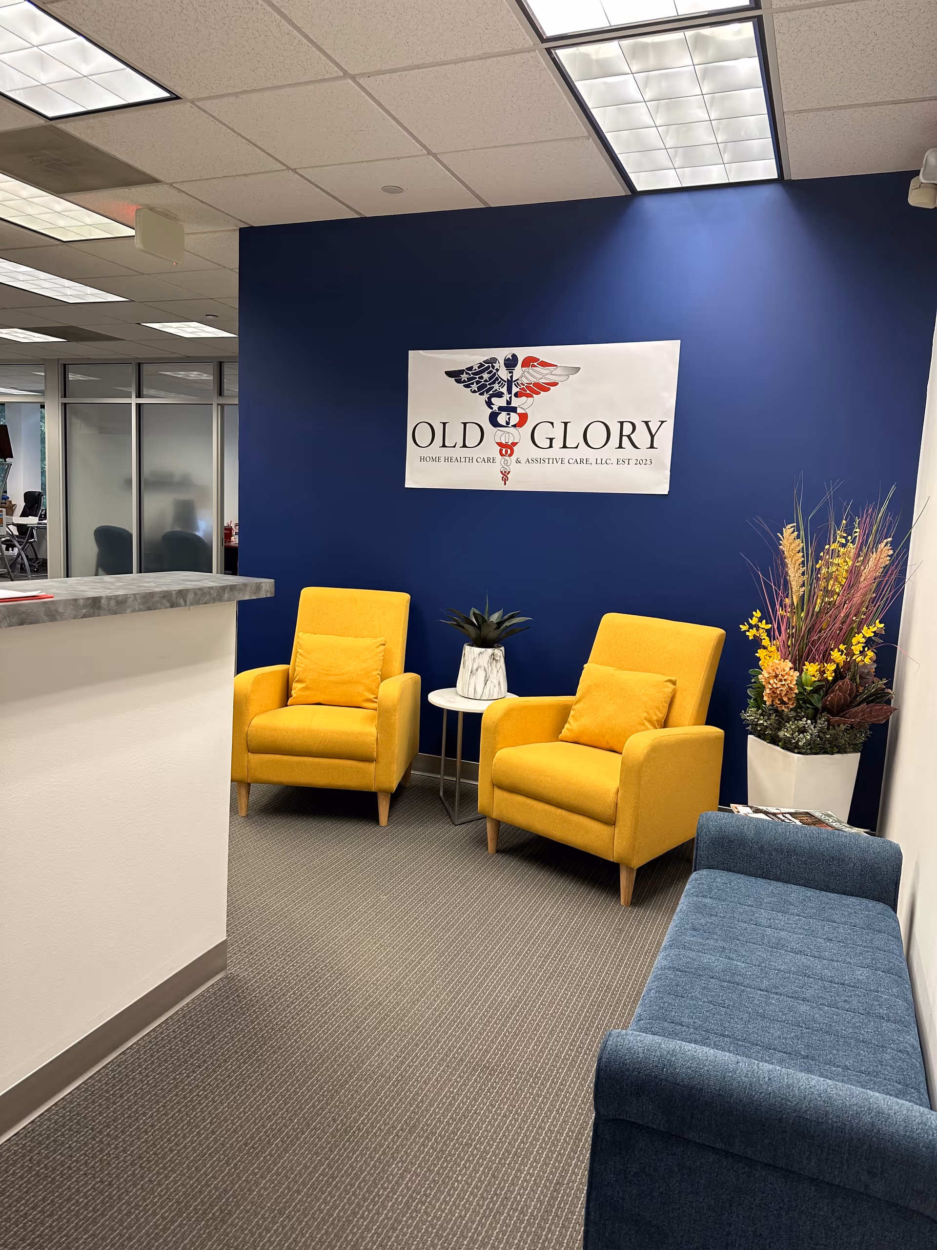 Reception lounge with blue wall, yellow chairs, plant, and Old Glory sign.
