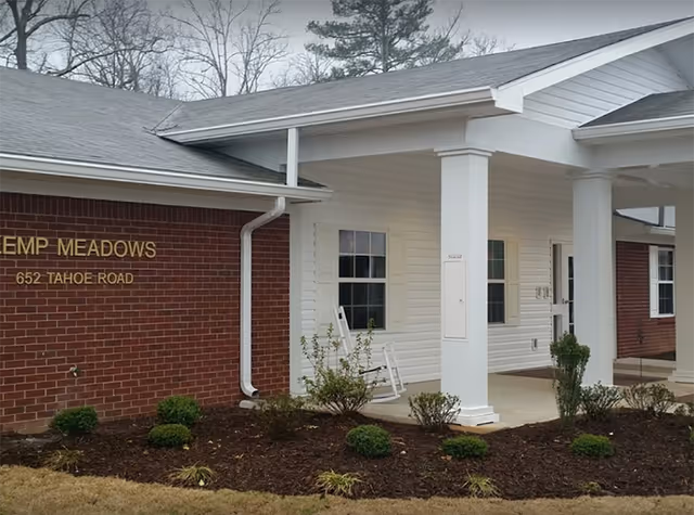 Exterior view of Kemp Meadows Assisted Living facility showing a brick and white siding building with a covered entrance supported by white columns, landscaping with small bushes, and a white rocking chair on the porch.
