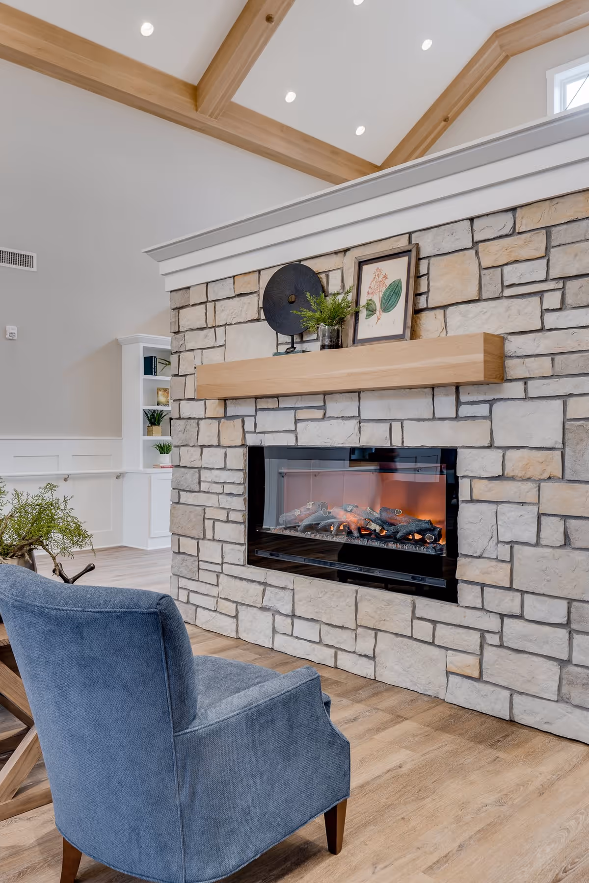 A cozy indoor living area featuring a stone fireplace with a wooden mantel. On the mantel are decorative items including a framed botanical print, a round black sculpture, and a small plant. In front of the fireplace is a comfortable blue upholstered armchair. The room has wooden flooring, white walls, and exposed wooden beams on the ceiling with recessed lighting.