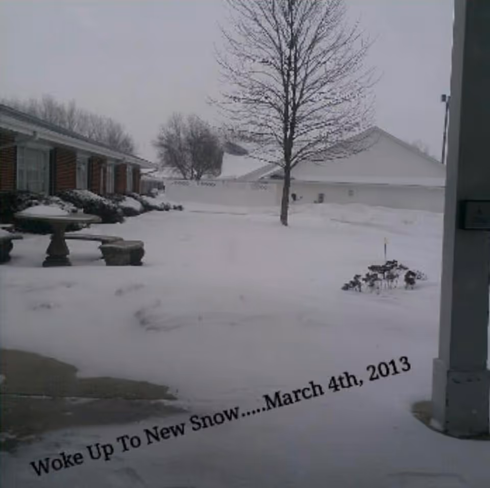 Snow blankets the senior living campus exterior, with a bare tree and nearby brick buildings.