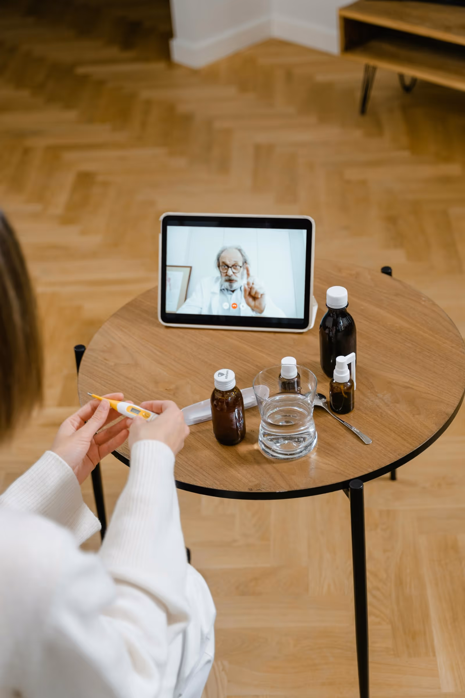 A tablet on a table shows a doctor in a video call, with medicine bottles nearby.