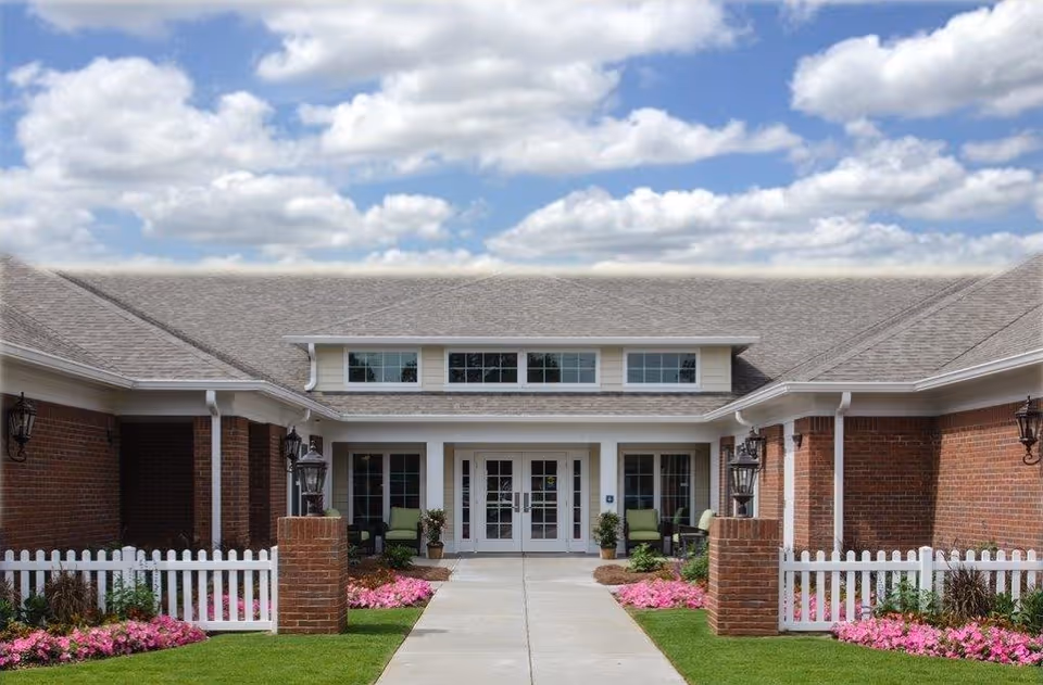 Front exterior view of a single-story brick building with a gray shingled roof, white trim, and a white picket fence. The entrance features double glass doors flanked by windows, with green chairs and potted plants on either side. Pink flowers and green grass line the walkway leading to the entrance under a partly cloudy sky.