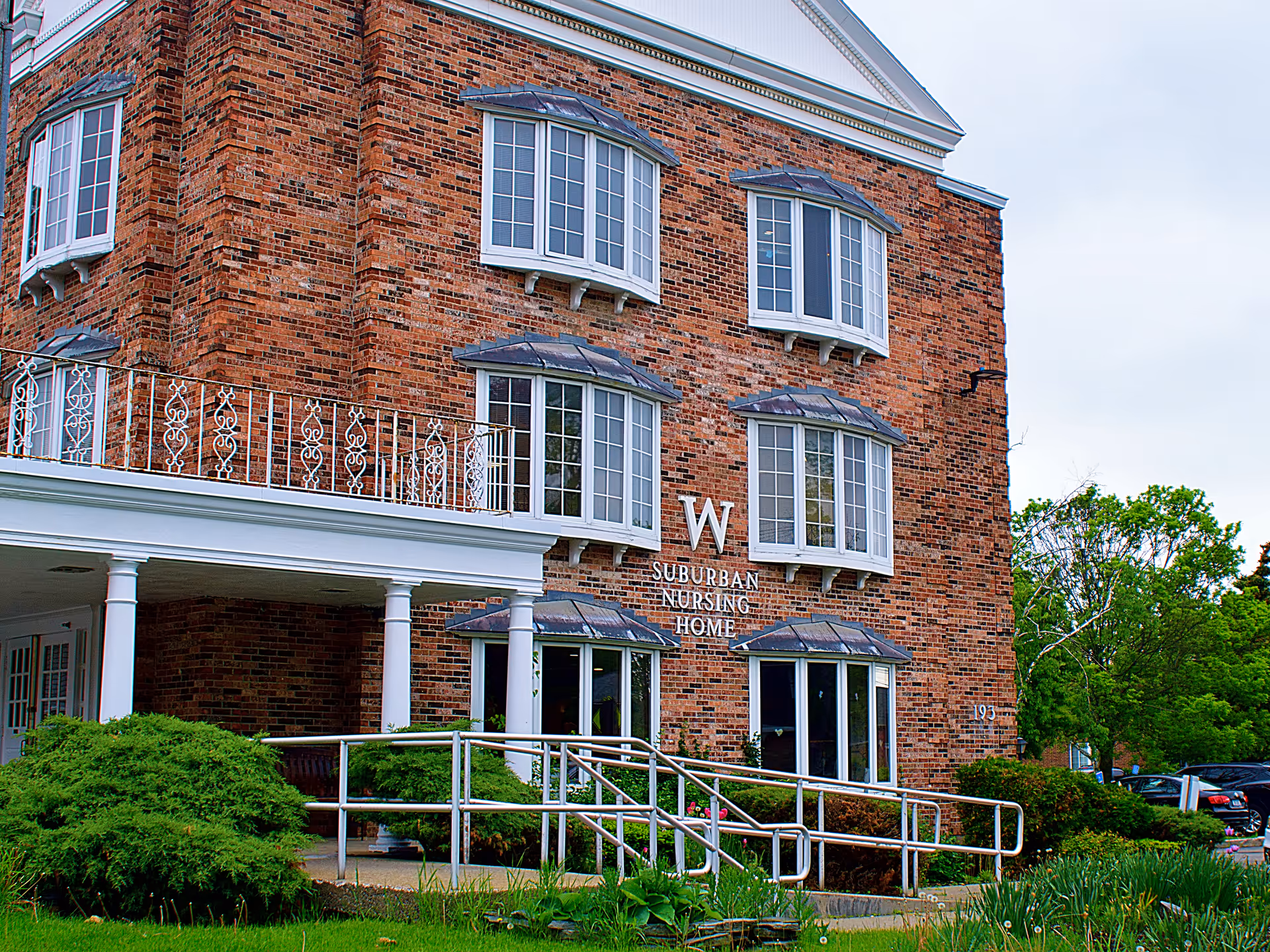 Brick nursing home exterior with white columns, balconies, and a ramp entrance.