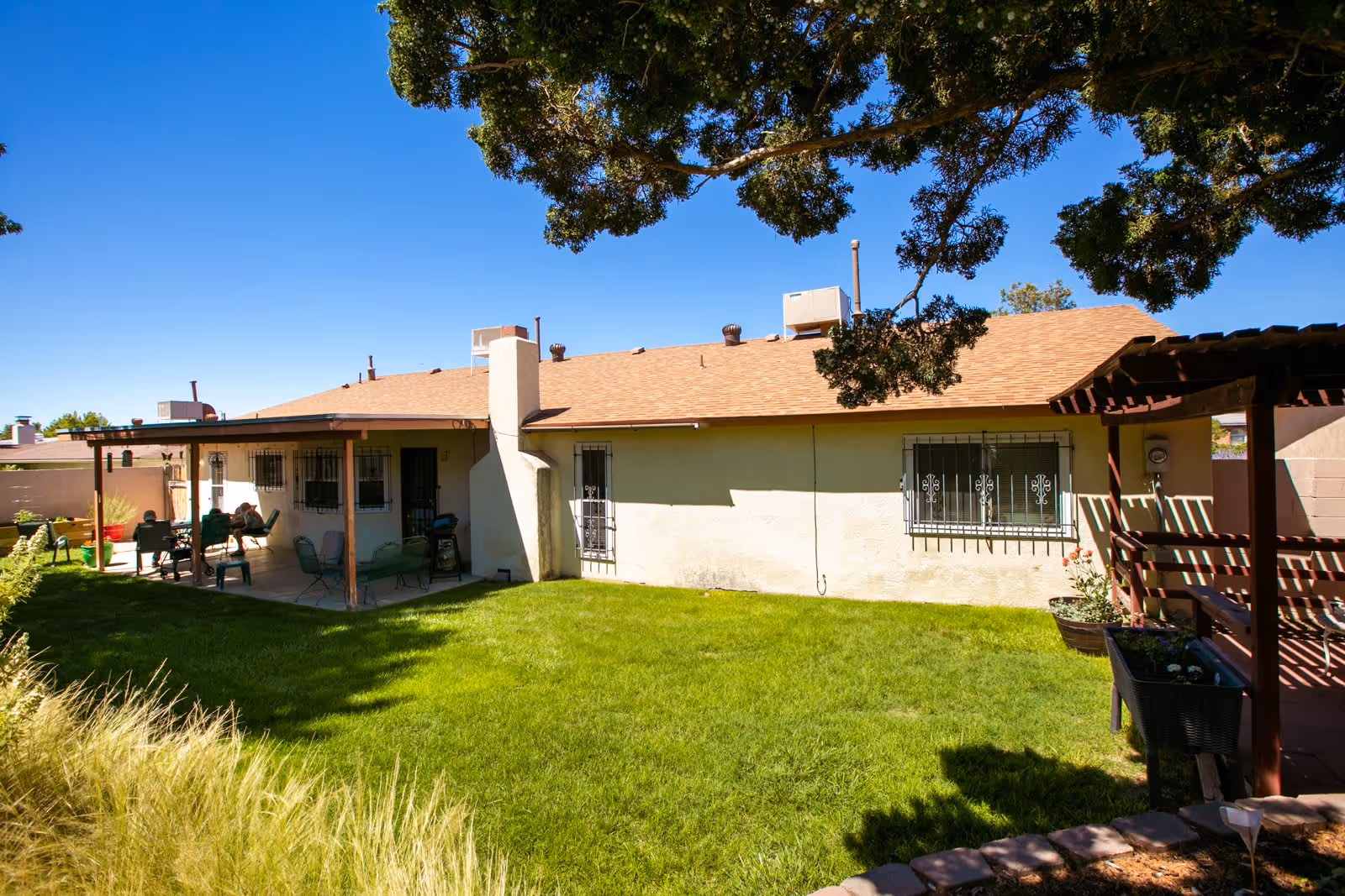 A sunny backyard area of a single-story building with a covered patio featuring outdoor seating. The yard has green grass, a large tree providing shade, and a wooden pergola with planters. The building has a light-colored exterior with barred windows and a chimney.