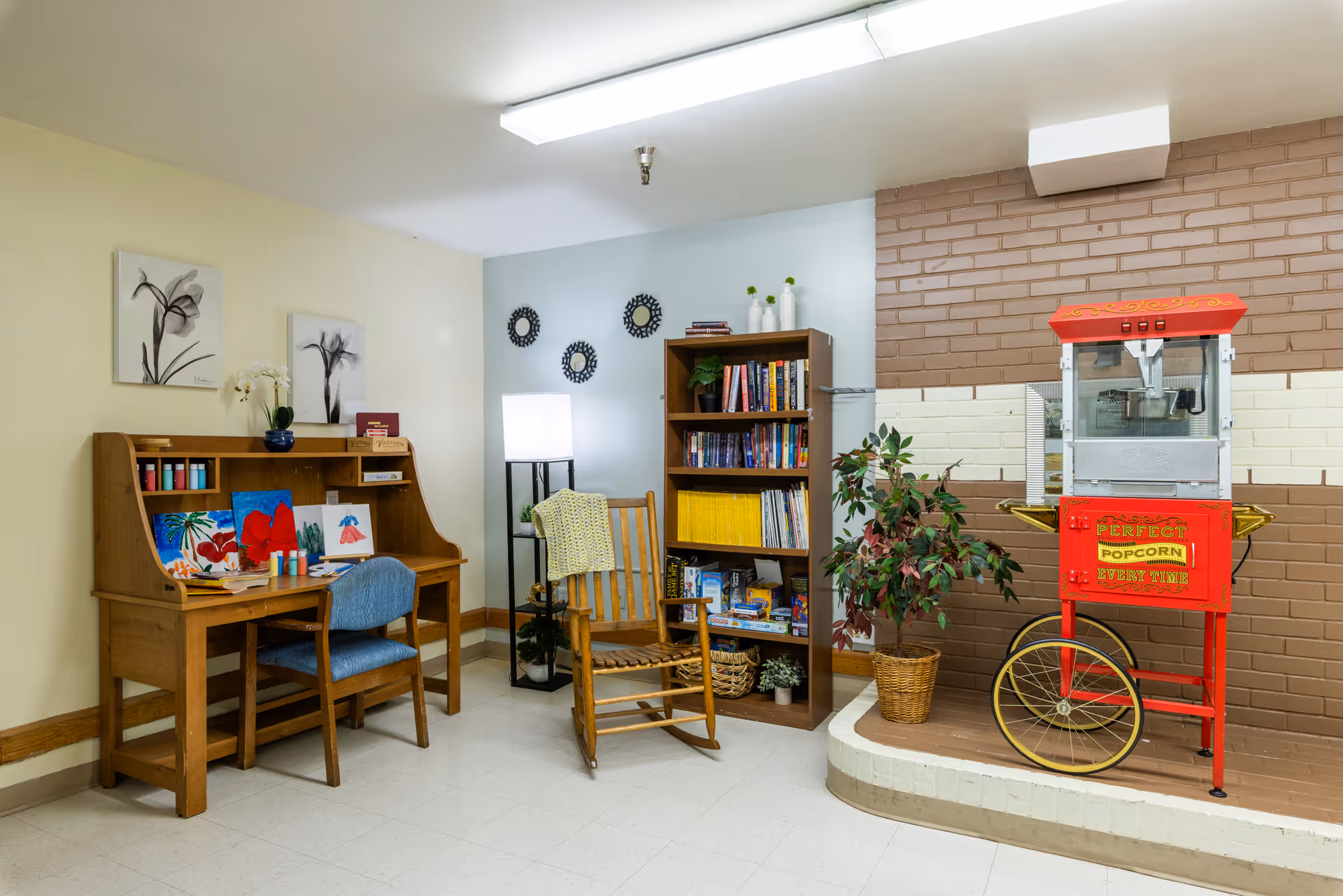 Interior common area with a wooden desk, bookshelf, rocking chair, plants, and a popcorn machine.
