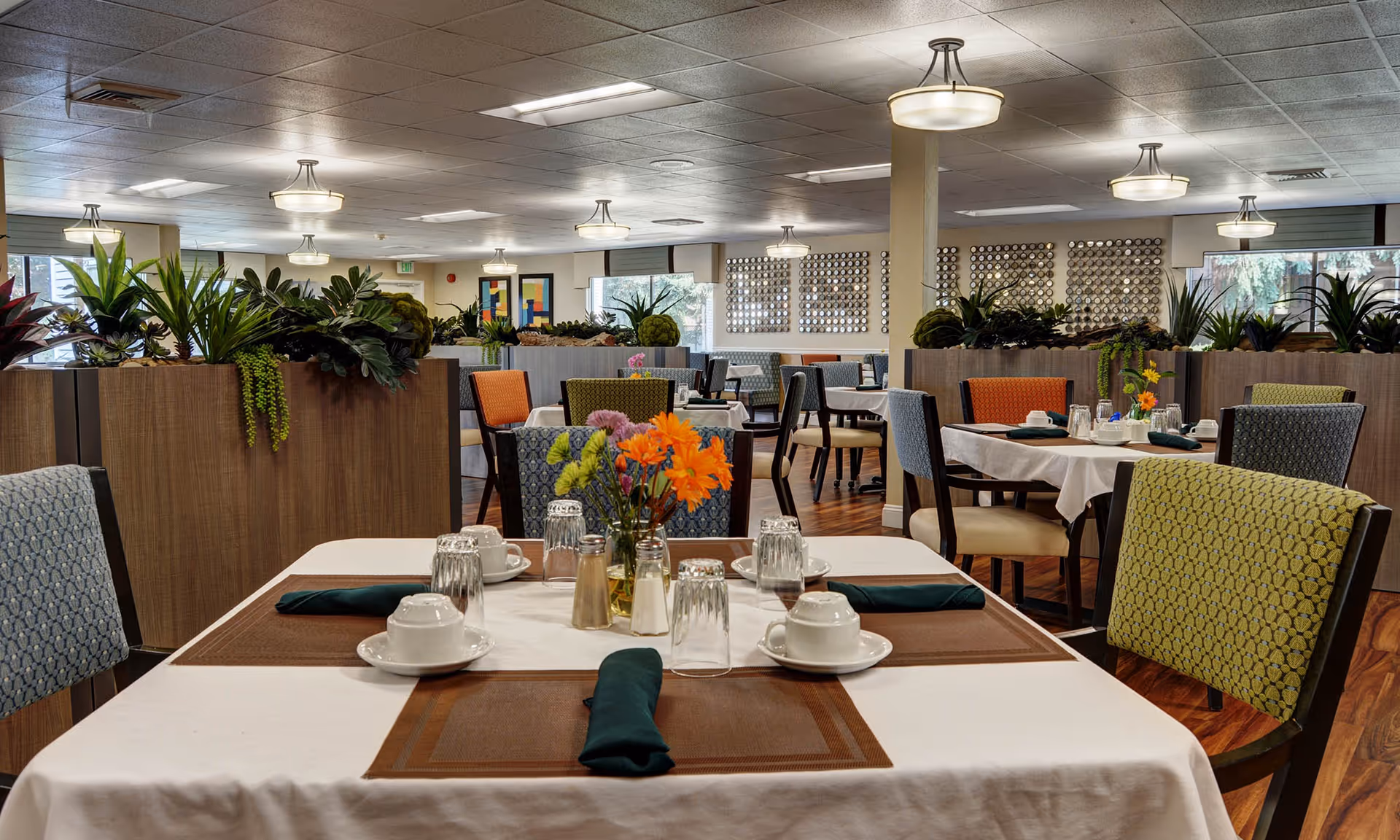 A spacious and well-lit dining room in a senior living facility with multiple tables set with white tablecloths, brown placemats, cups, glasses, and green napkins. The chairs have colorful patterned upholstery in shades of blue, orange, and green. There are decorative plants on wooden dividers separating the seating areas, and the room features ceiling lights and large windows allowing natural light.