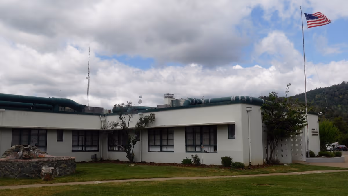 White low-slung building with exposed pipes, a tall flagpole, and a grassy lawn.