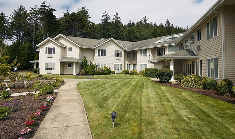Exterior view of a two-story assisted living residence with a central walkway, manicured lawn, and landscaped gardens.