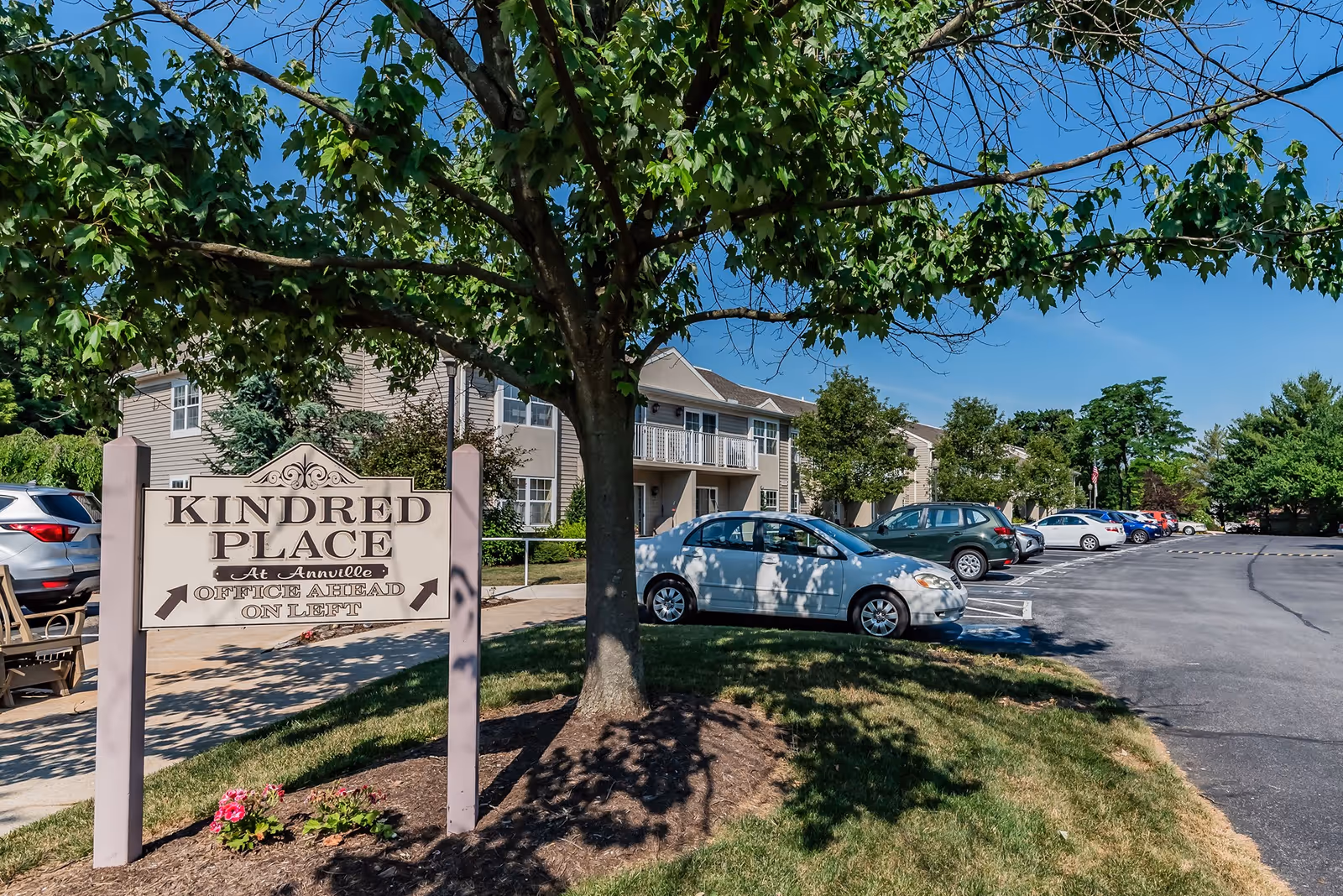 Outdoor view of Kindred Place At Annville showing a parking lot with several parked cars, a large tree in the foreground, and a sign indicating the office is ahead on the left. Residential buildings are visible in the background under a clear blue sky.