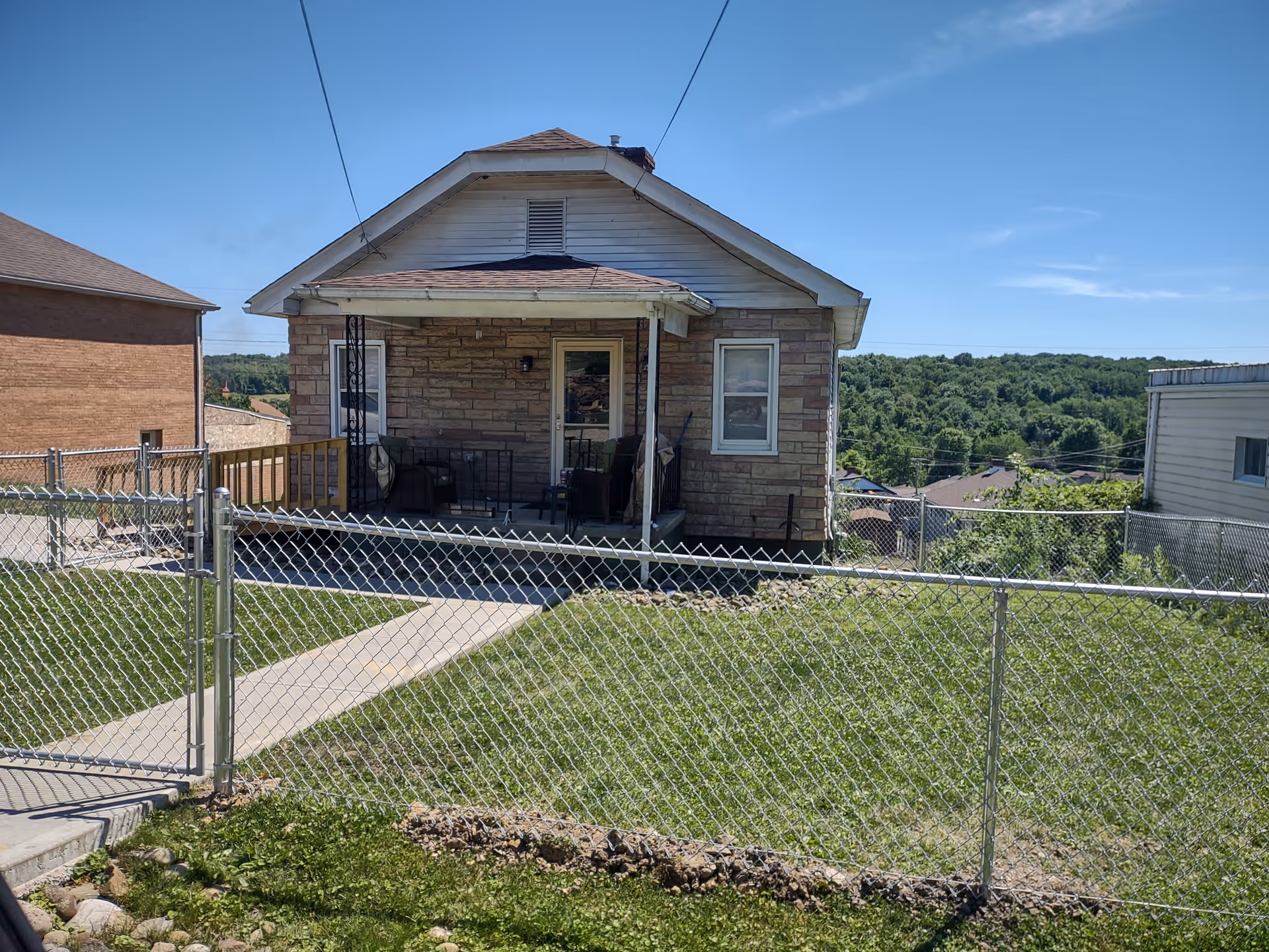 Small brick-front building with a porch, chain-link fence, and grassy yard on a sunny day.