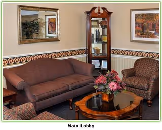 A cozy lobby area with a brown upholstered sofa, two patterned armchairs, a wooden coffee table with a glass top and a floral centerpiece. Behind the sofa is a large framed mirror, and to the right is a wooden display cabinet with decorative items. A framed painting hangs on the wall above one armchair.
