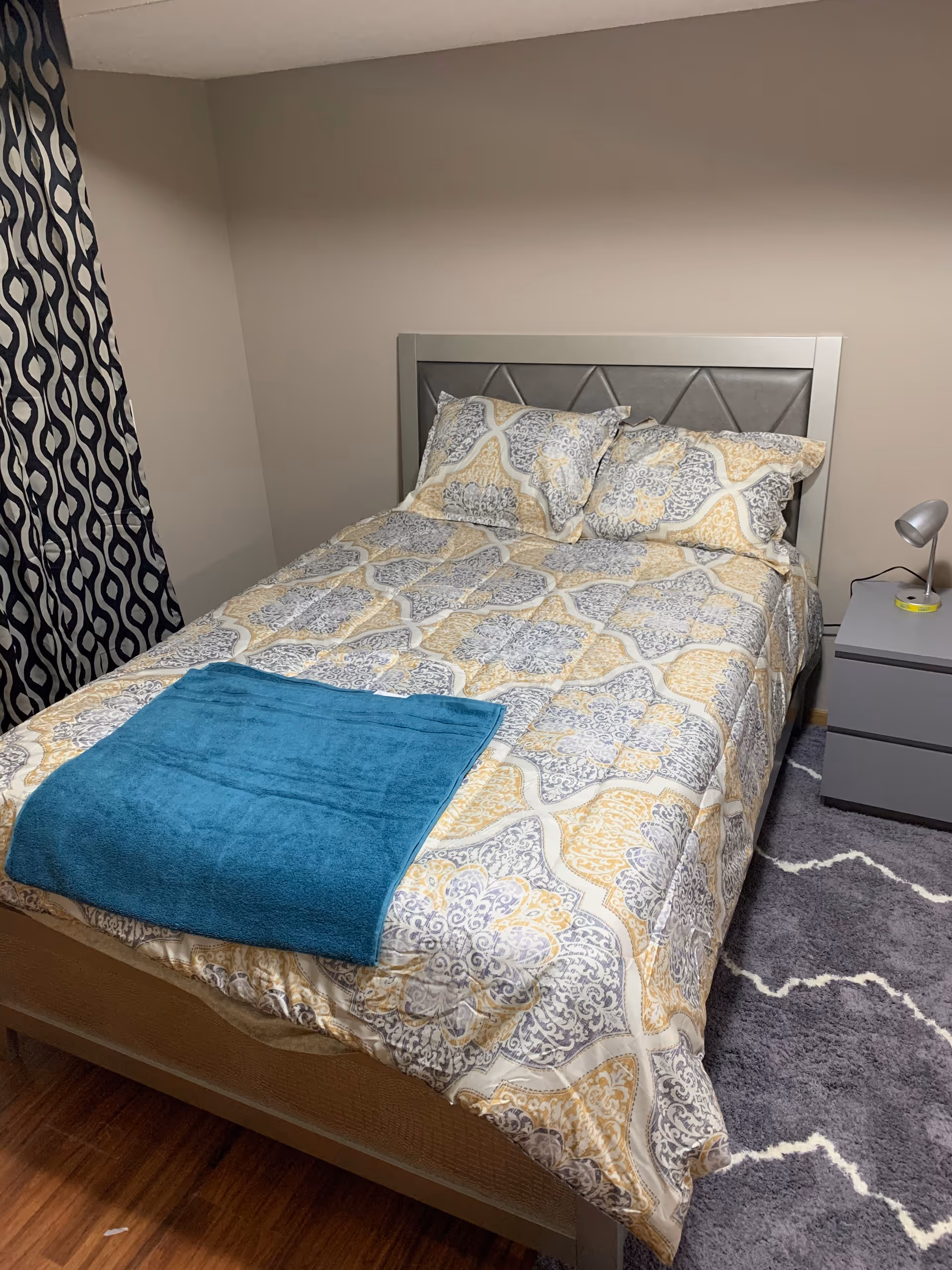 A neatly made bed with a patterned bedspread in shades of gray, yellow, and white, with two matching pillows and a folded blue towel at the foot of the bed. Next to the bed is a gray nightstand with a small silver lamp and a yellow item on it. The room has beige walls, a patterned curtain in black and white, and a gray rug with a white zigzag pattern on a wooden floor.