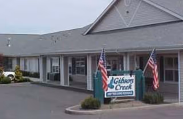 Exterior view of a single-story building with a gray roof and light-colored siding. There are two American flags on either side of a sign that reads 'Gibson Creek Memory Care.' The building has multiple windows and a covered walkway in front.