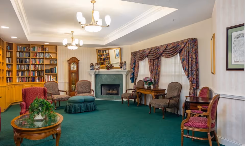 Community sitting room with bookshelves, a fireplace, multiple upholstered chairs and tables on green carpet.