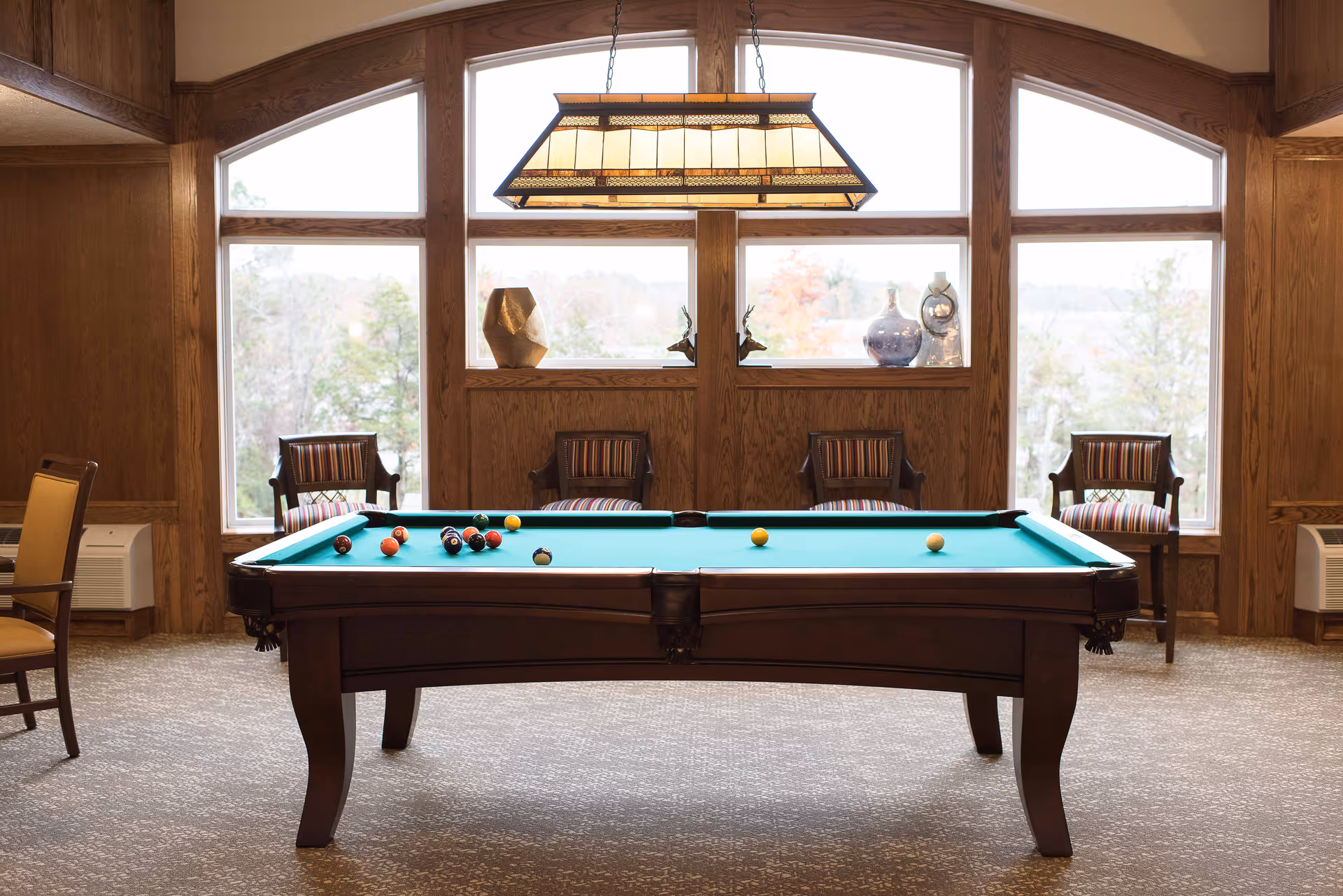 A pool table with billiard balls set up in a room with large windows and wooden paneling. Four wooden chairs with striped cushions are placed against the wall under the windows, and a rectangular hanging light fixture is above the pool table.