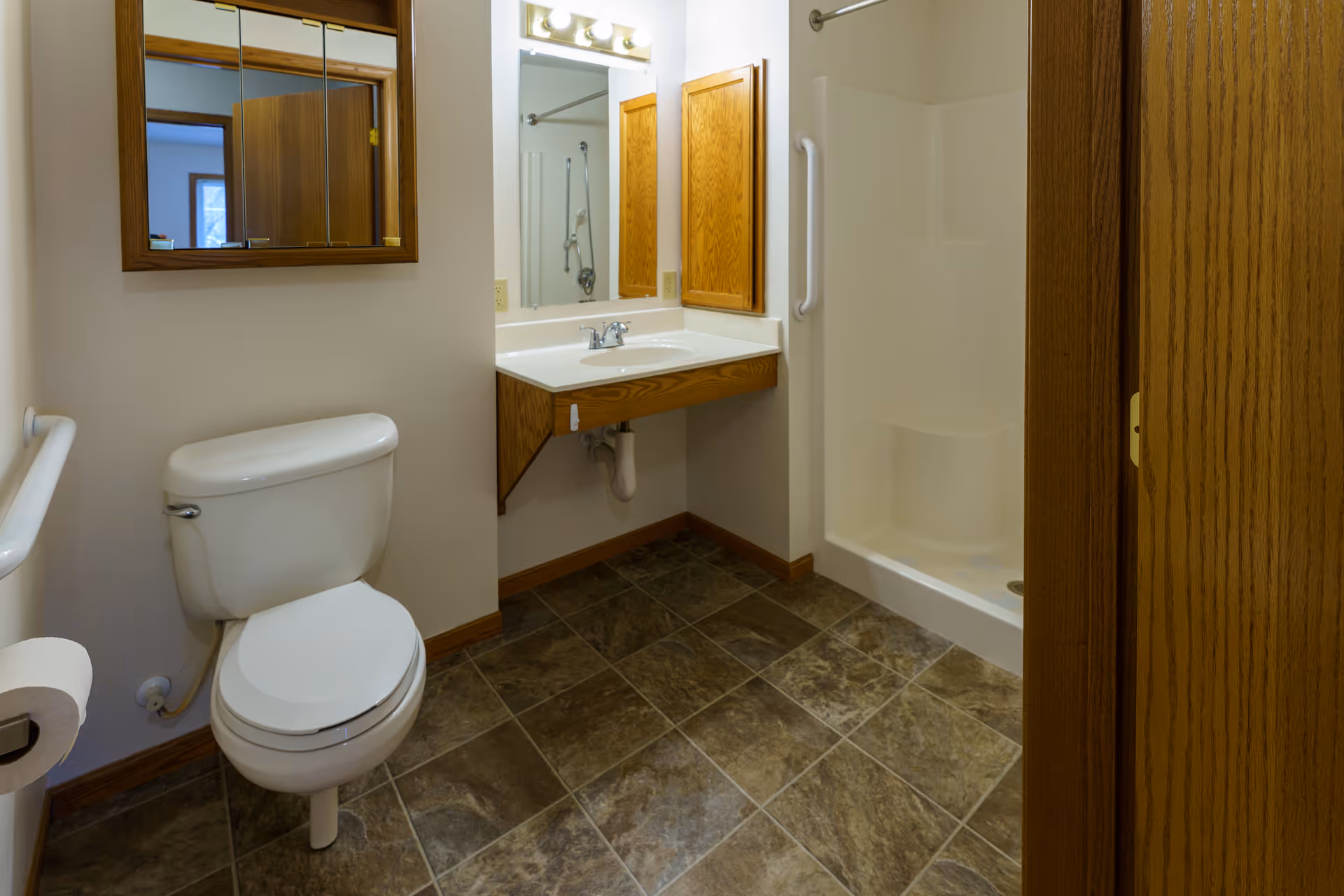 A clean bathroom with a white toilet, a wall-mounted sink with a wooden cabinet above it, a large mirror with lights above, and a walk-in shower with a built-in seat and grab bar. The floor is covered with brown and gray tiles, and the walls are painted white.