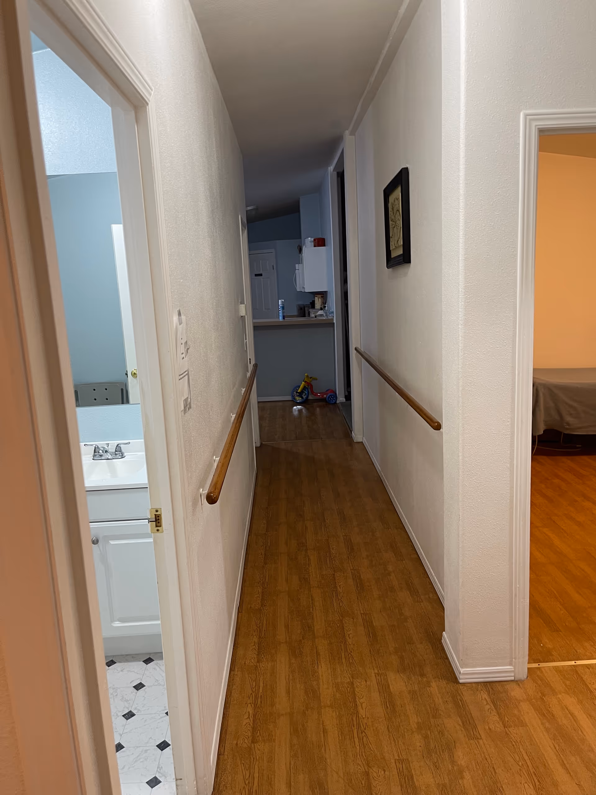 Narrow hallway inside a senior living facility with wood floors, bathroom doorway, kitchen at end, handrails.