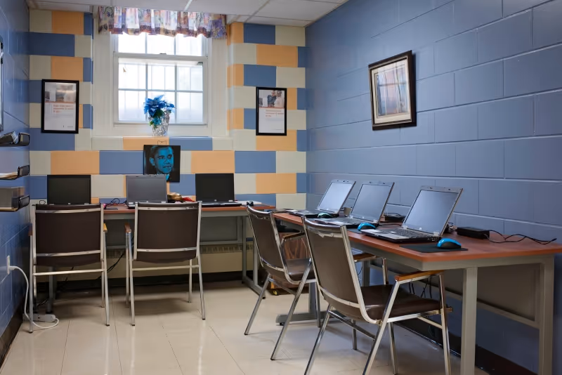 Small computer room in a senior facility with multiple laptops and chairs along two tables beneath a window and colorful block-painted walls.