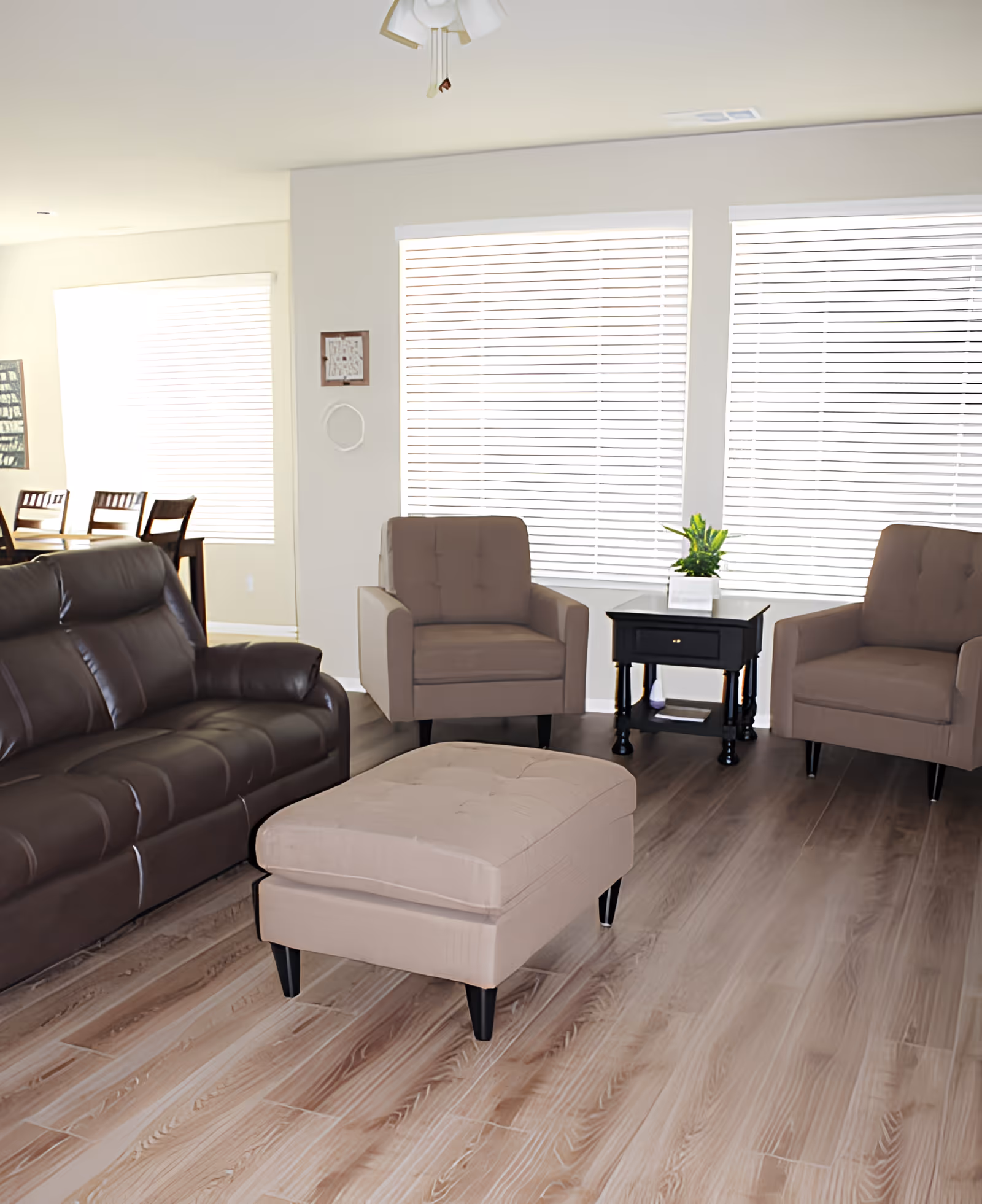 A bright living room with a dark brown leather sofa, two beige armchairs, a matching beige ottoman, and a black side table with a small green plant on top. The room has large windows with white blinds and light wood flooring.