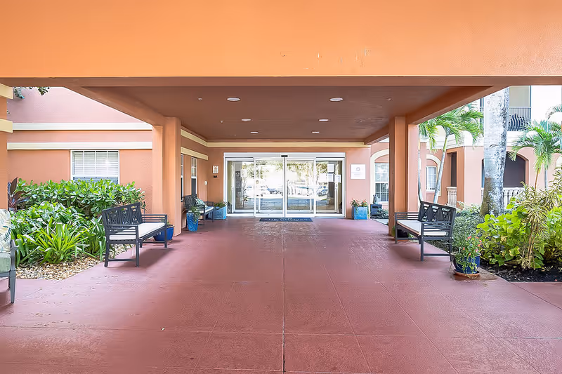 Covered entrance of a coral-colored building with sliding glass doors, benches, and potted plants.