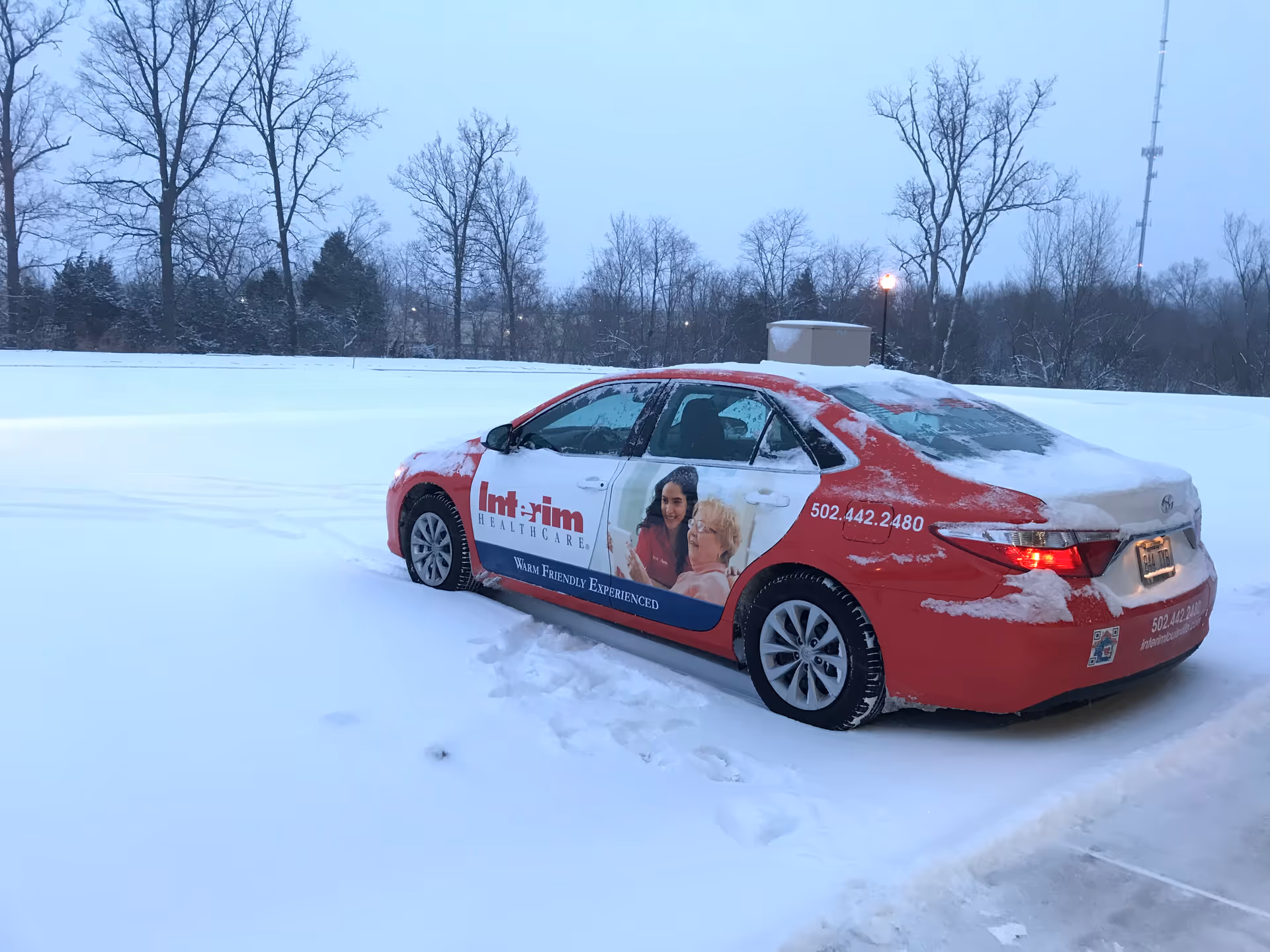 Red Interim HealthCare car parked on a snowy lot with bare trees in the background.
