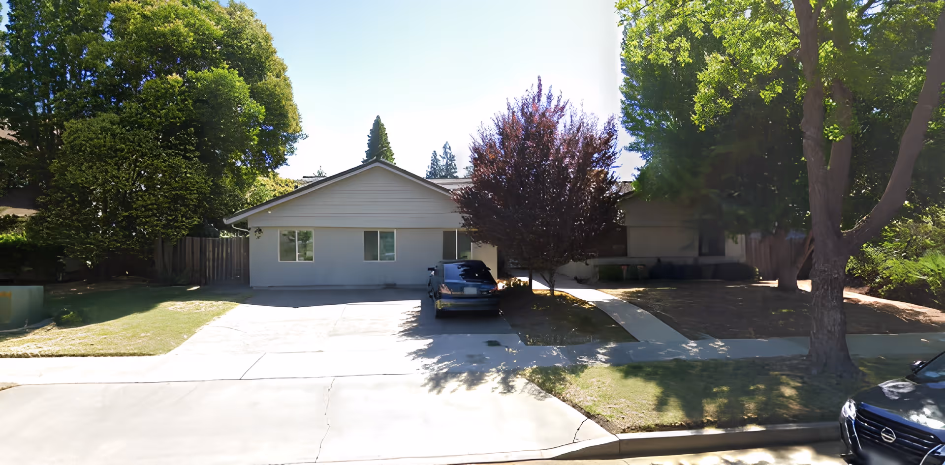 Single-story light-gray building with a driveway and a parked car, surrounded by trees and a front lawn.