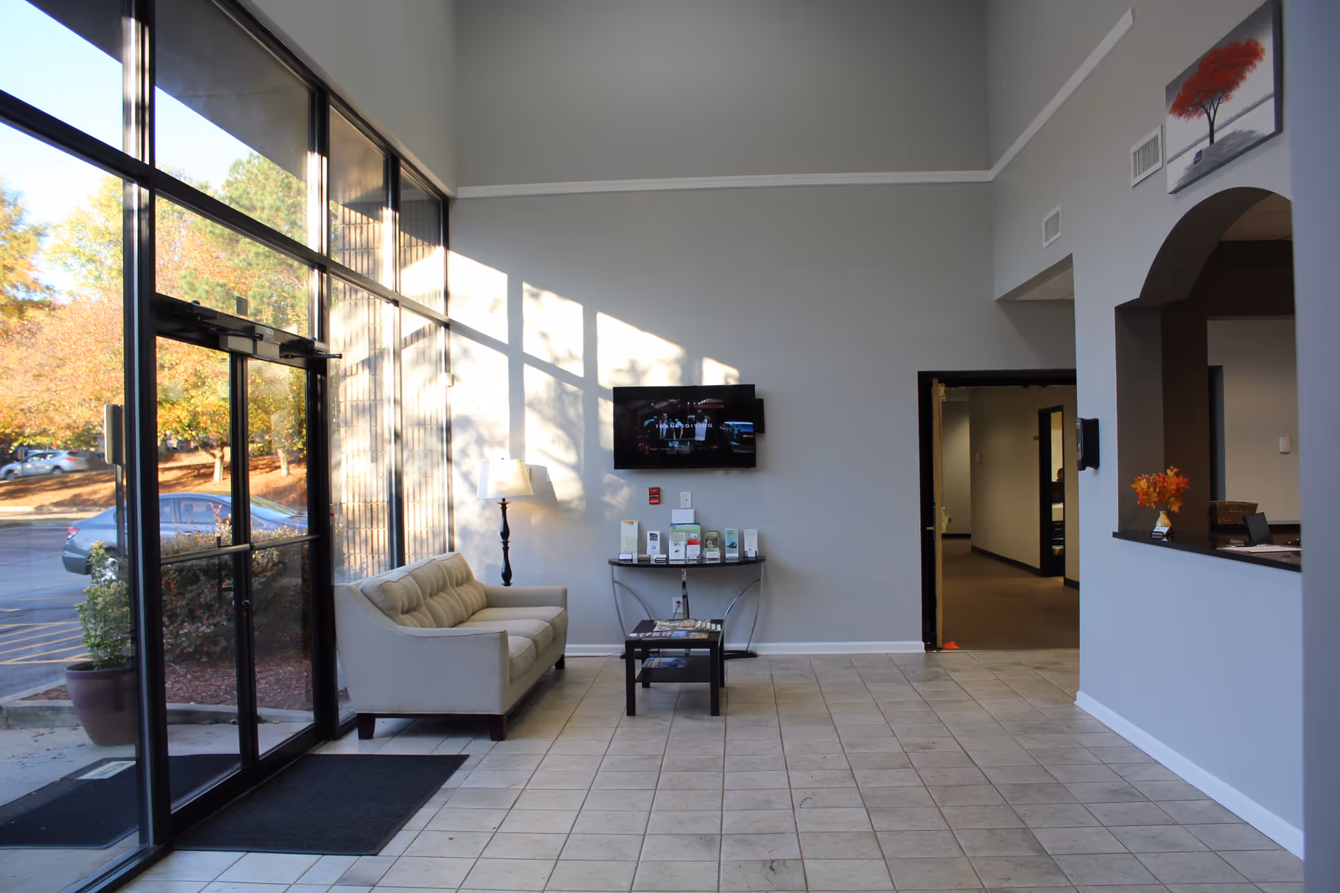 Bright lobby with glass doors, beige sofa, TV, and brochure table.