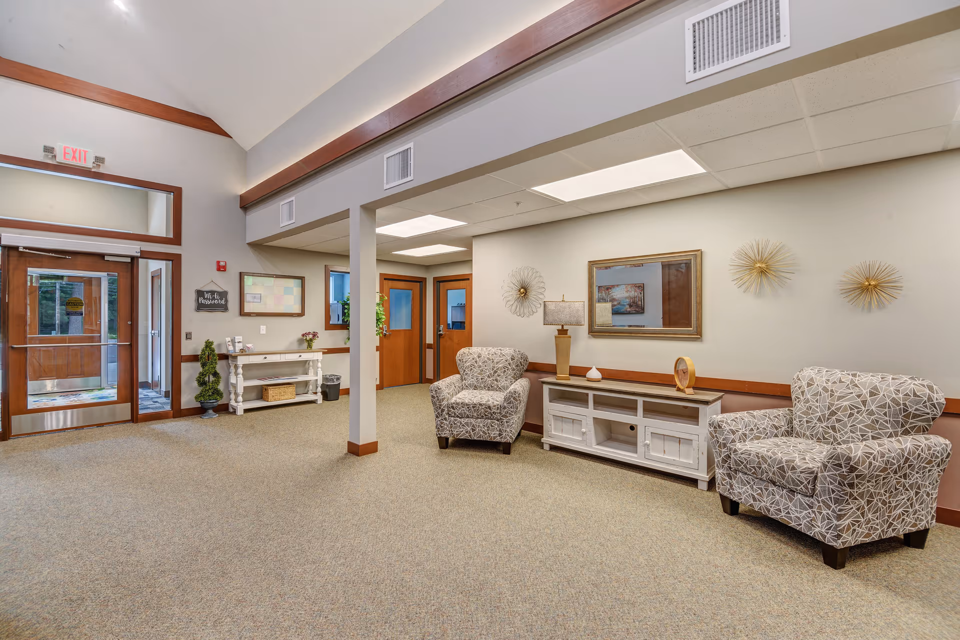 A bright and spacious senior living facility common area with two patterned armchairs, a white console table with decorative items including a lamp and clock, wall art, and a wooden door entrance with an exit sign above. The room has beige walls, carpeted floor, and ceiling lights.