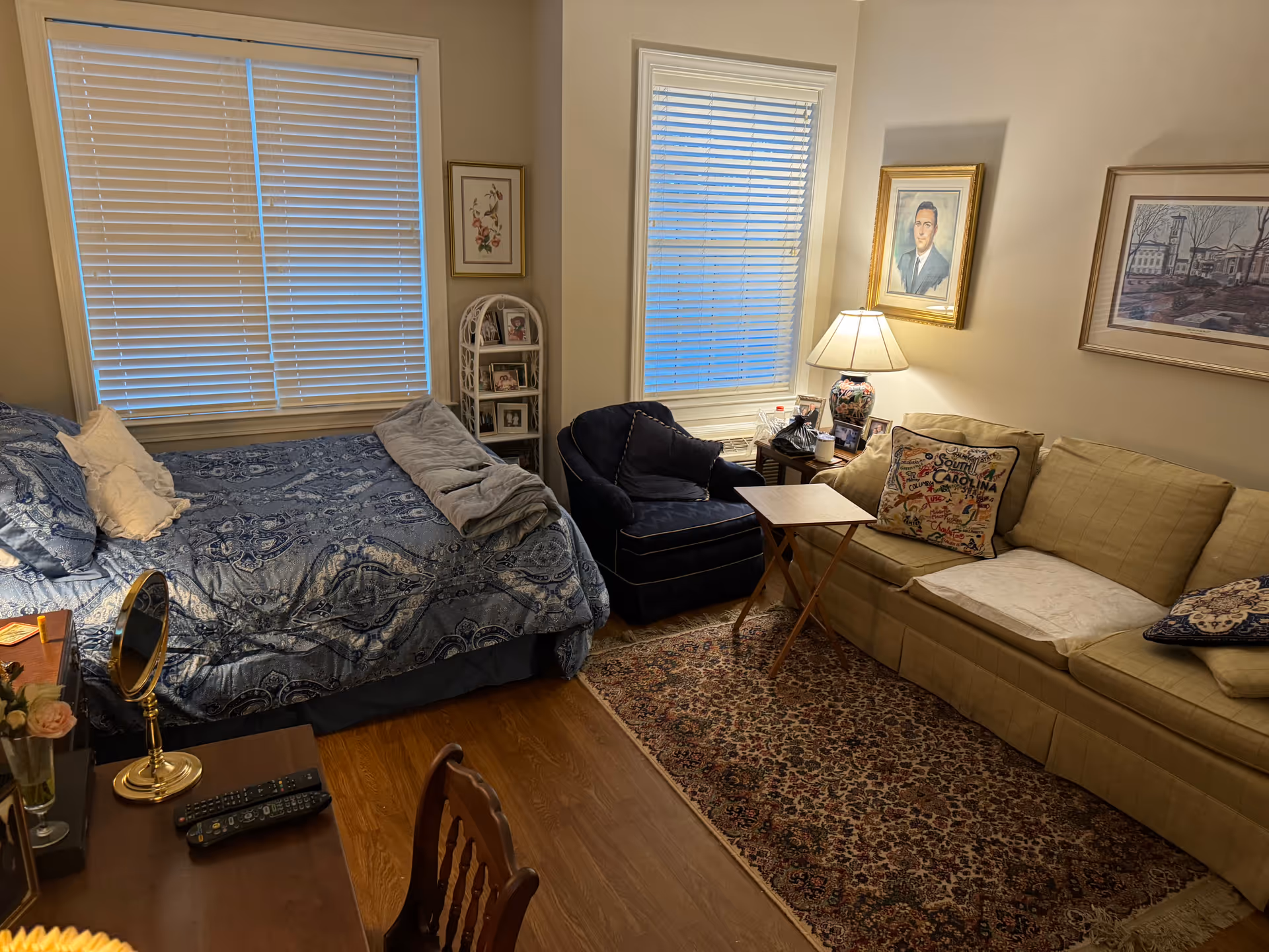 Cozy studio living area with a bed, beige sofa, blinds, and a patterned rug.