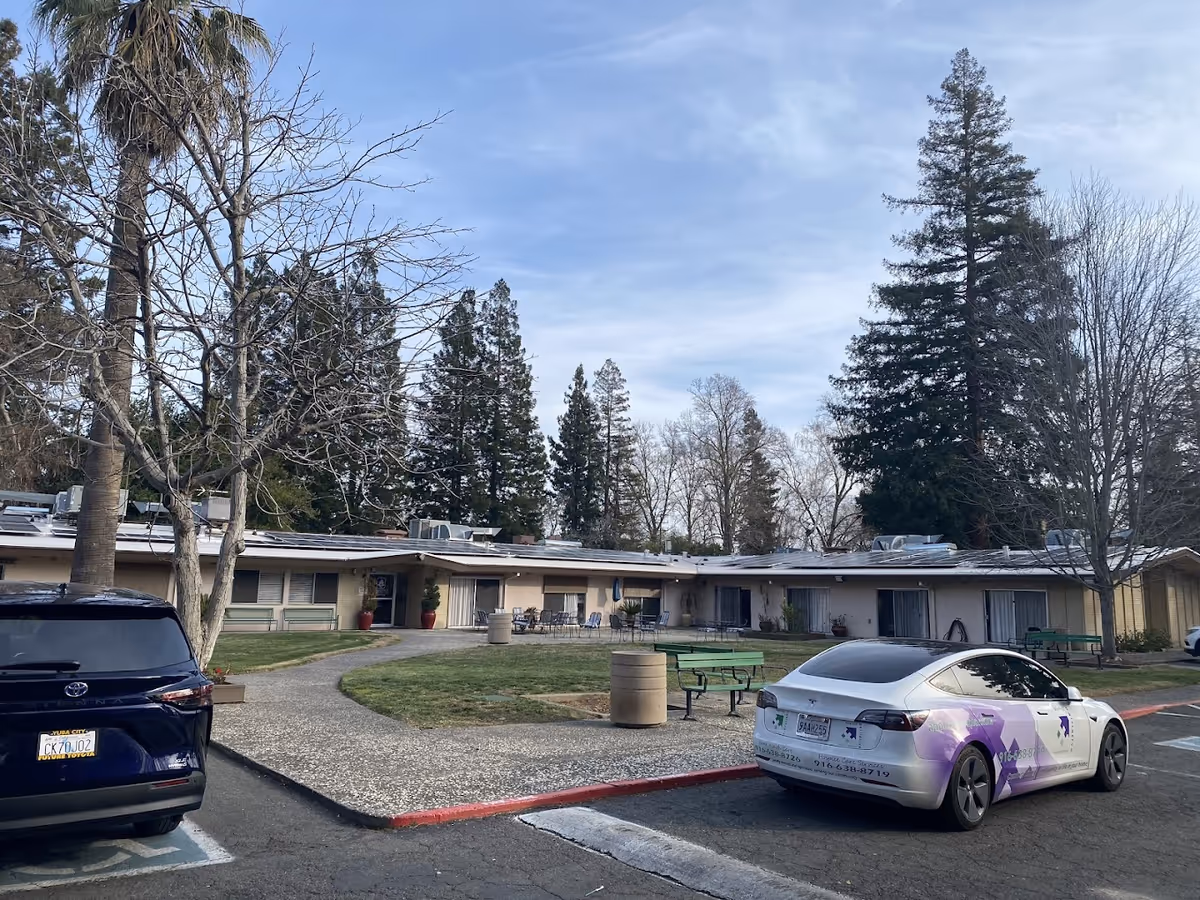 Single-story senior living building front with a small courtyard, benches, parked cars, and tall trees under a blue sky.